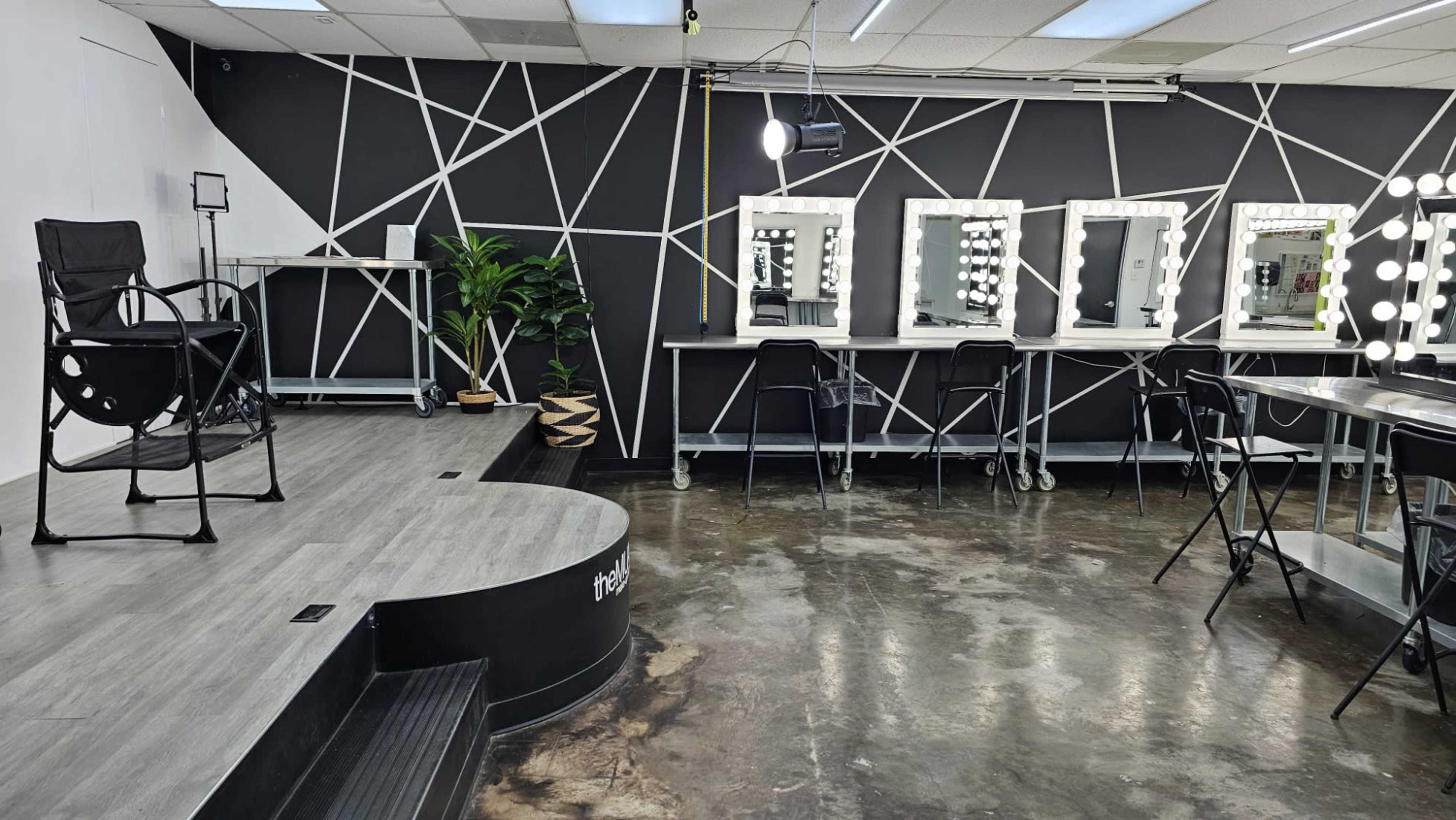 The image shows a modern makeup studio featuring a raised platform with a chair, a series of illuminated mirrors along one wall, and a sleek, black-and-white geometric design.