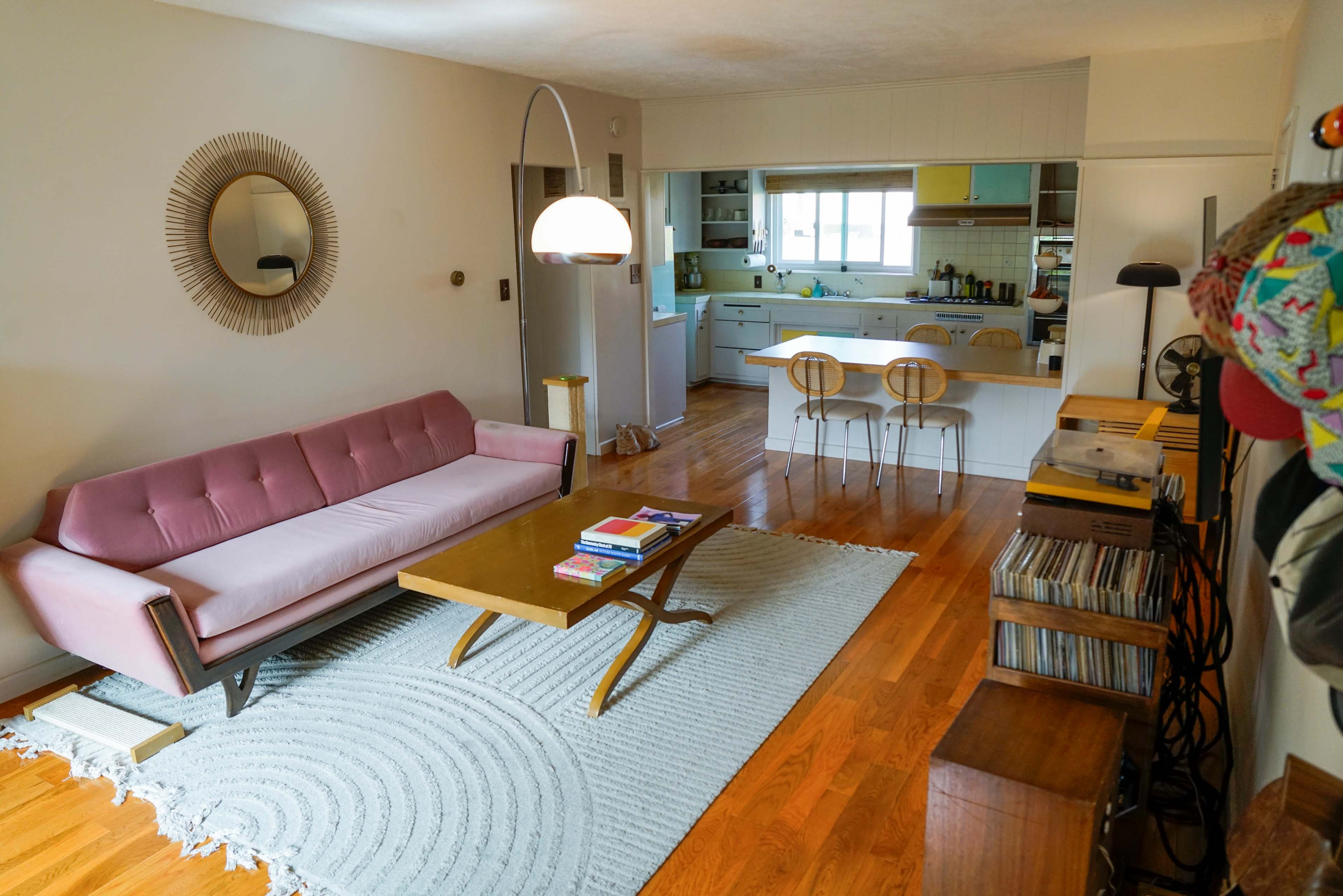 A pink sofa faces a wooden coffee table on a circular rug in a living room that opens into a kitchen area.