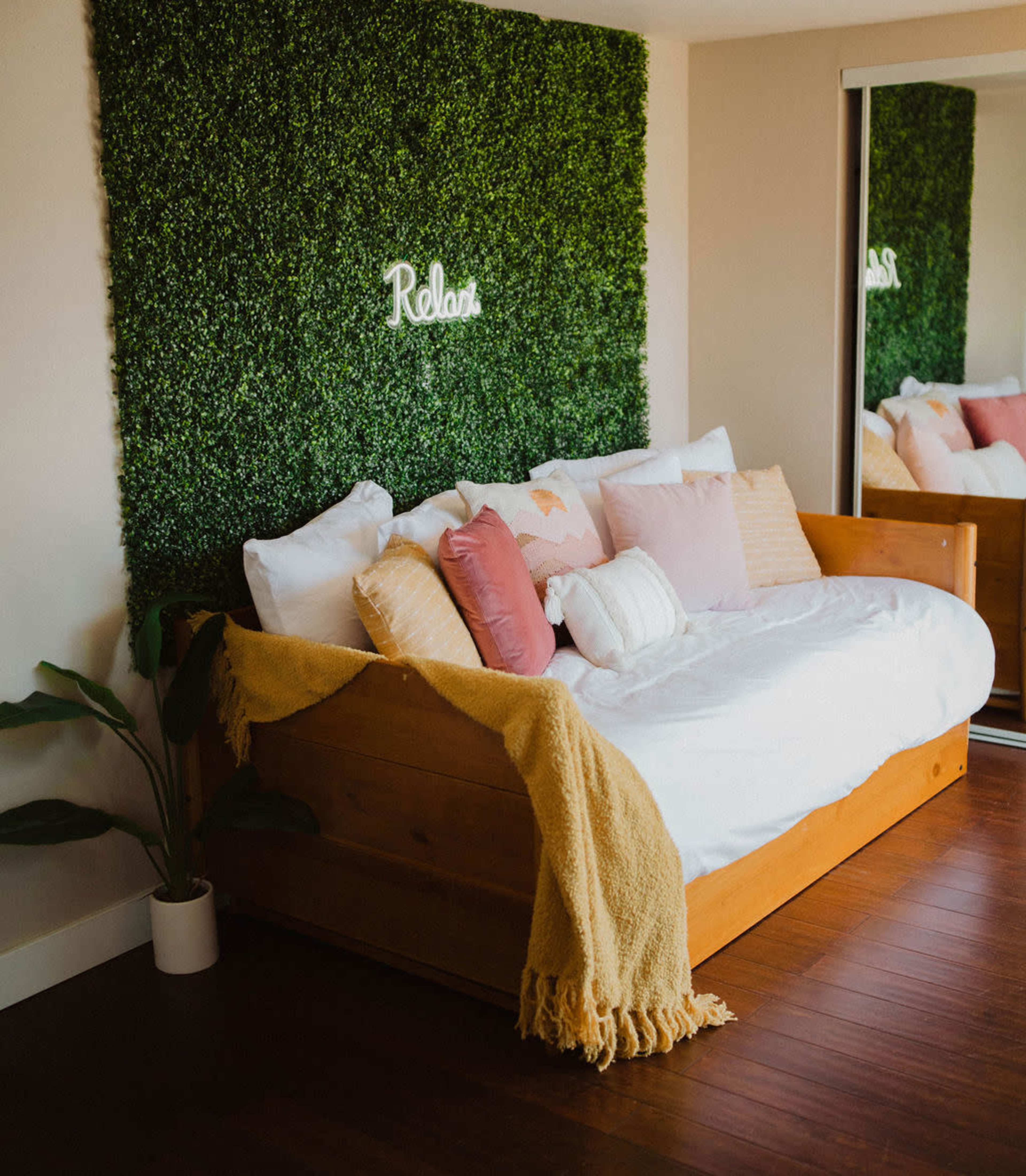 A cozy room features a wooden daybed with decorative pillows, a green hedge wall with the word "Relax" lit above it, and a mirror reflecting the space.
