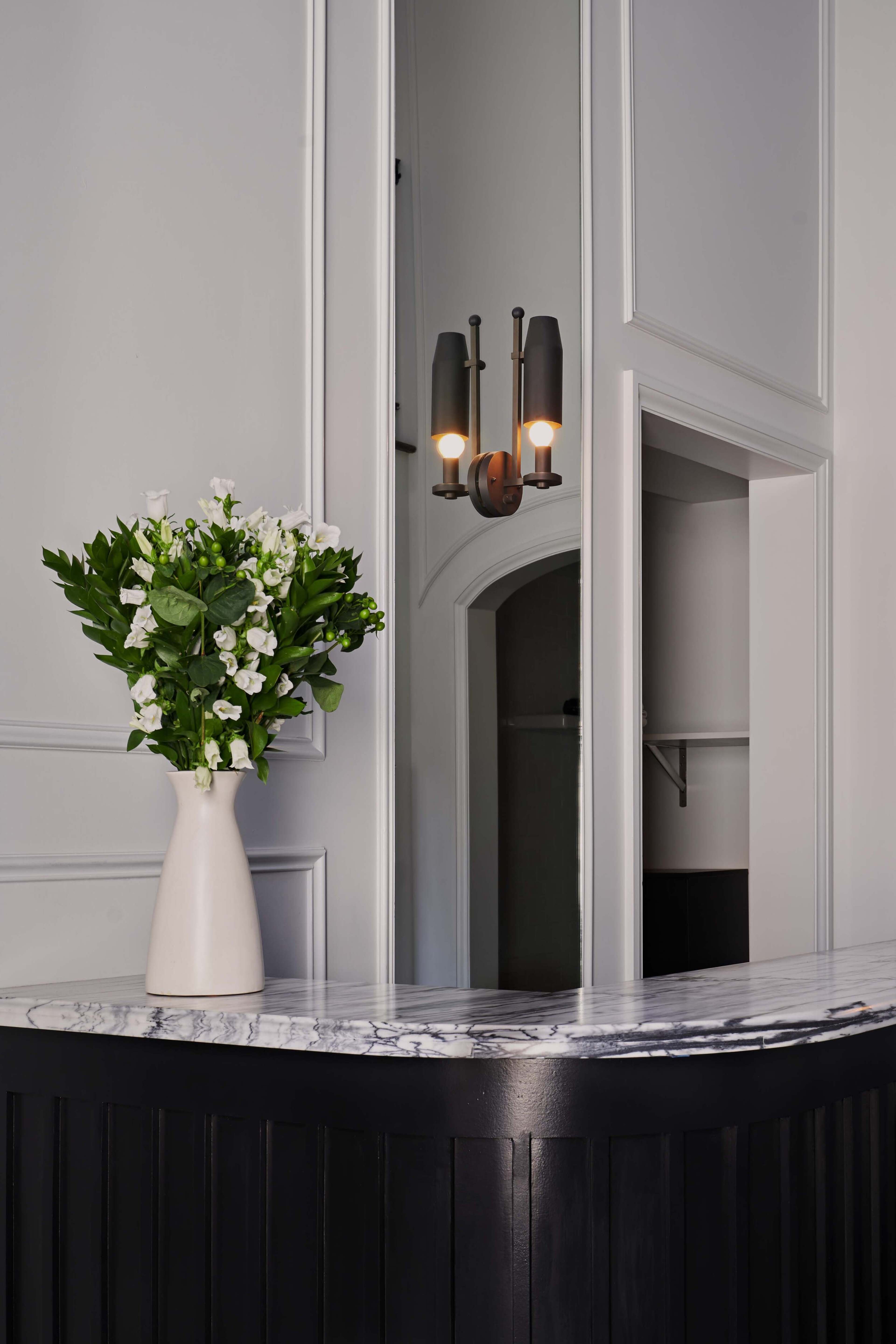 A vase of white flowers sits on a marble-topped counter beneath a mirror and a wall sconce in a simple interior space.