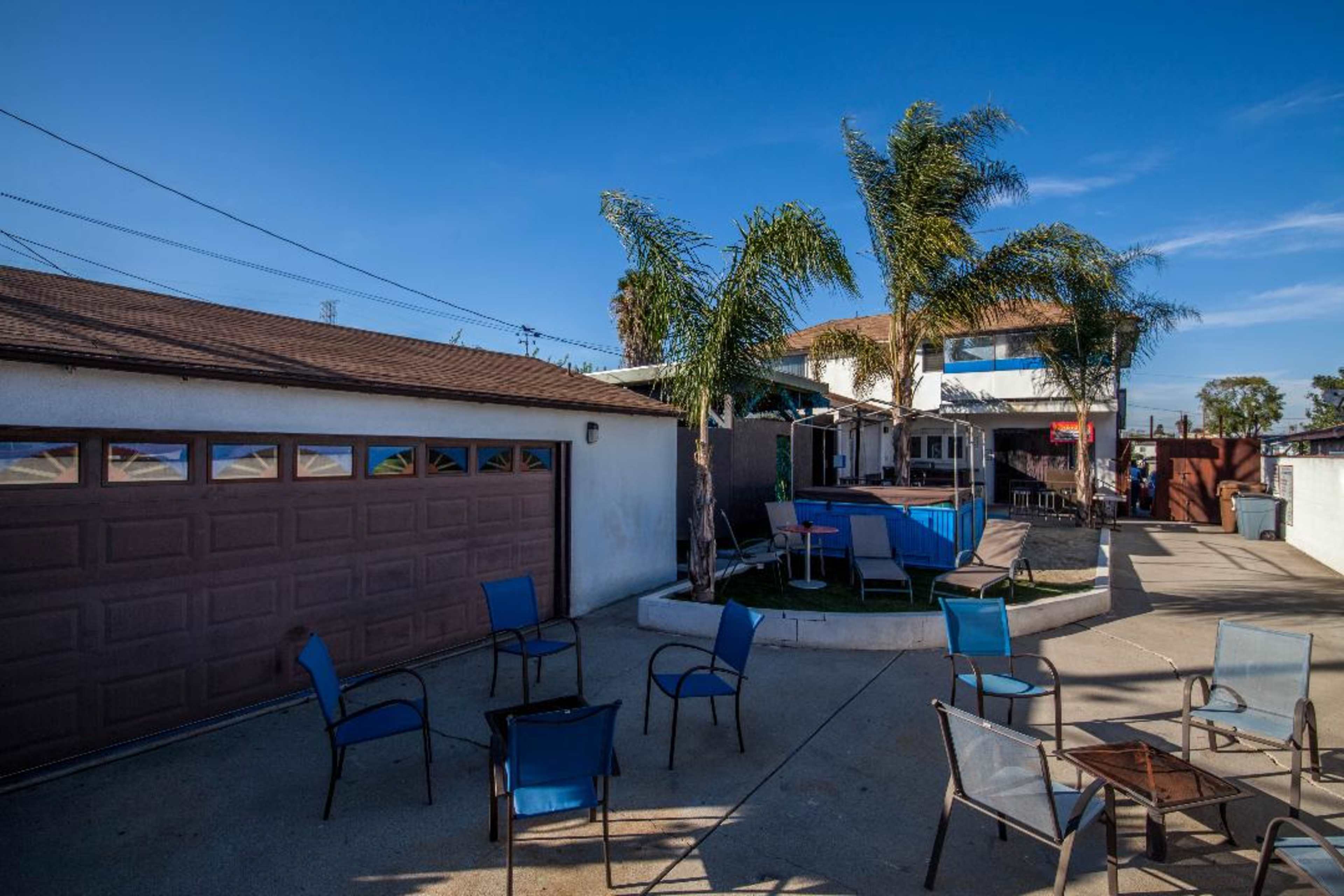 A patio area with several chairs is surrounded by palm trees and a house in the background.