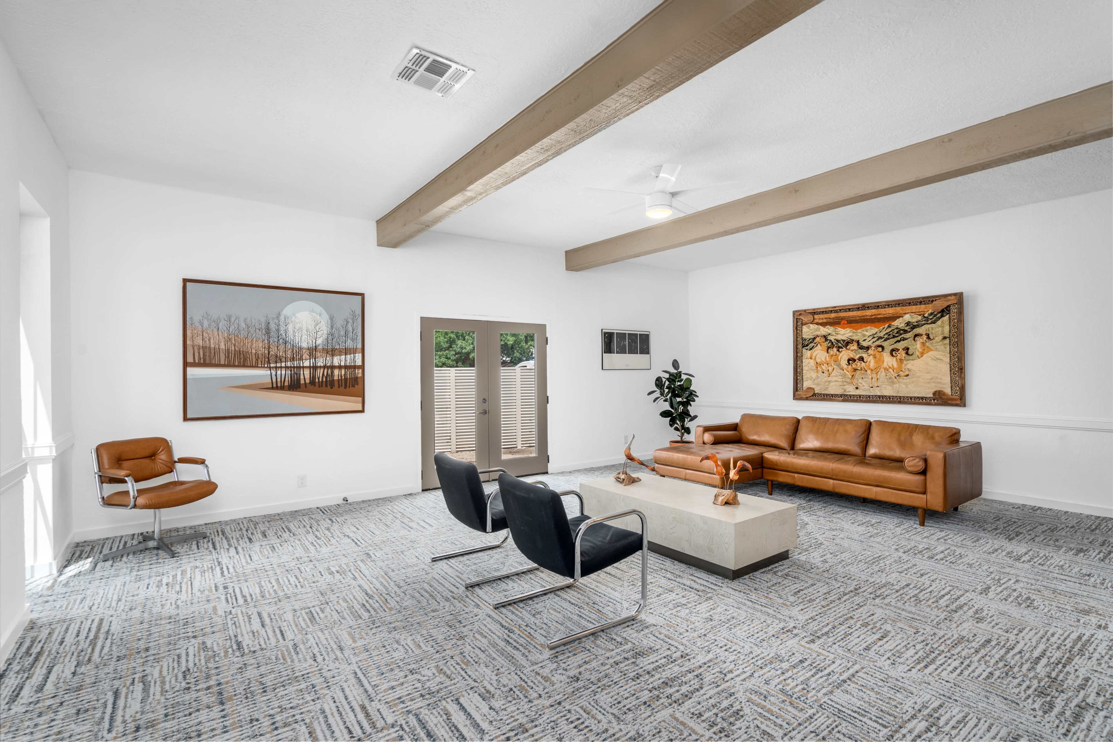 The image shows a spacious living room with a brown leather sofa, two black chairs, a coffee table, and artwork on the walls, all under a ceiling with exposed beams.