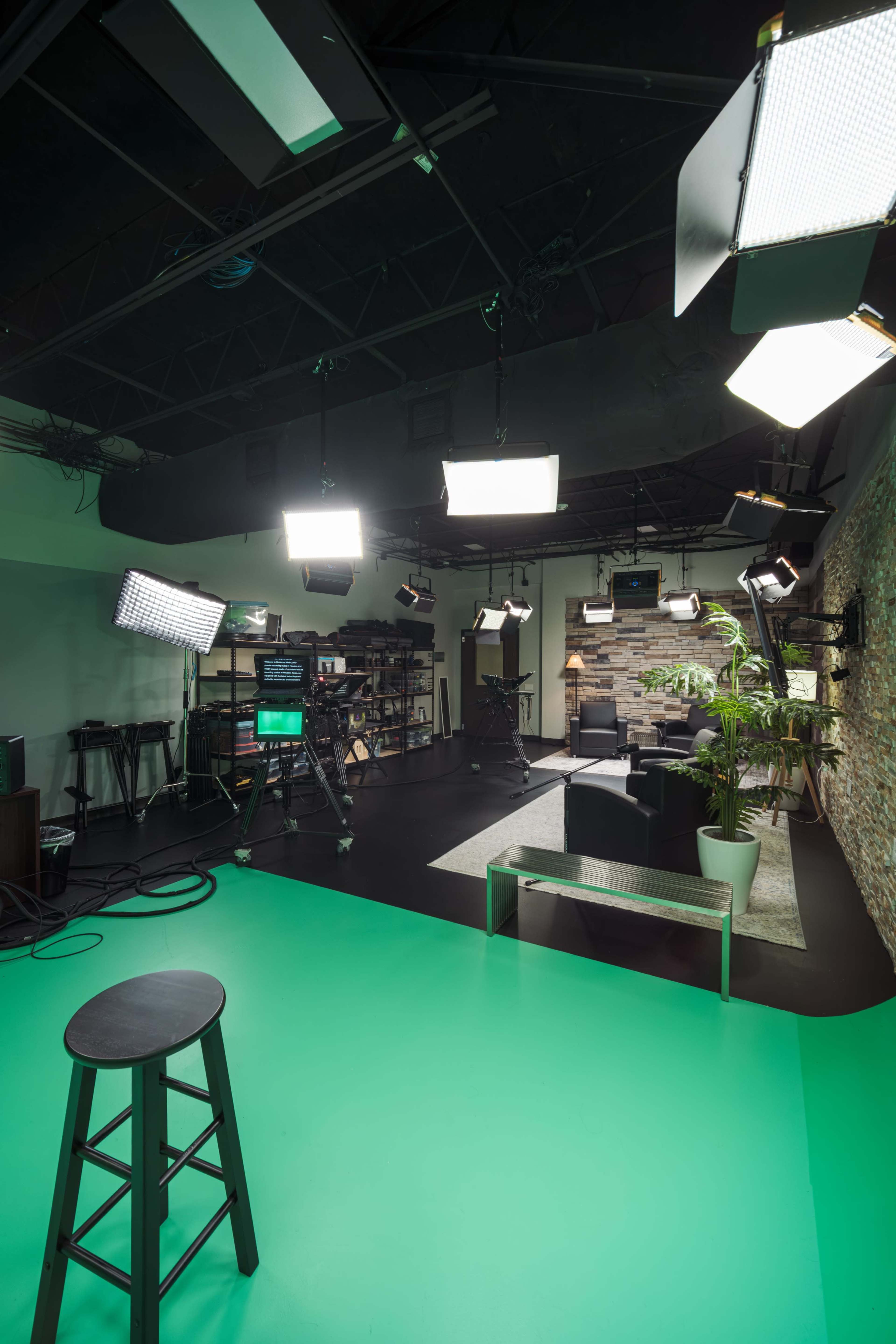 The image shows a television studio setup with a green floor, various lights, cameras, and a seating area featuring a black couch and a plant.