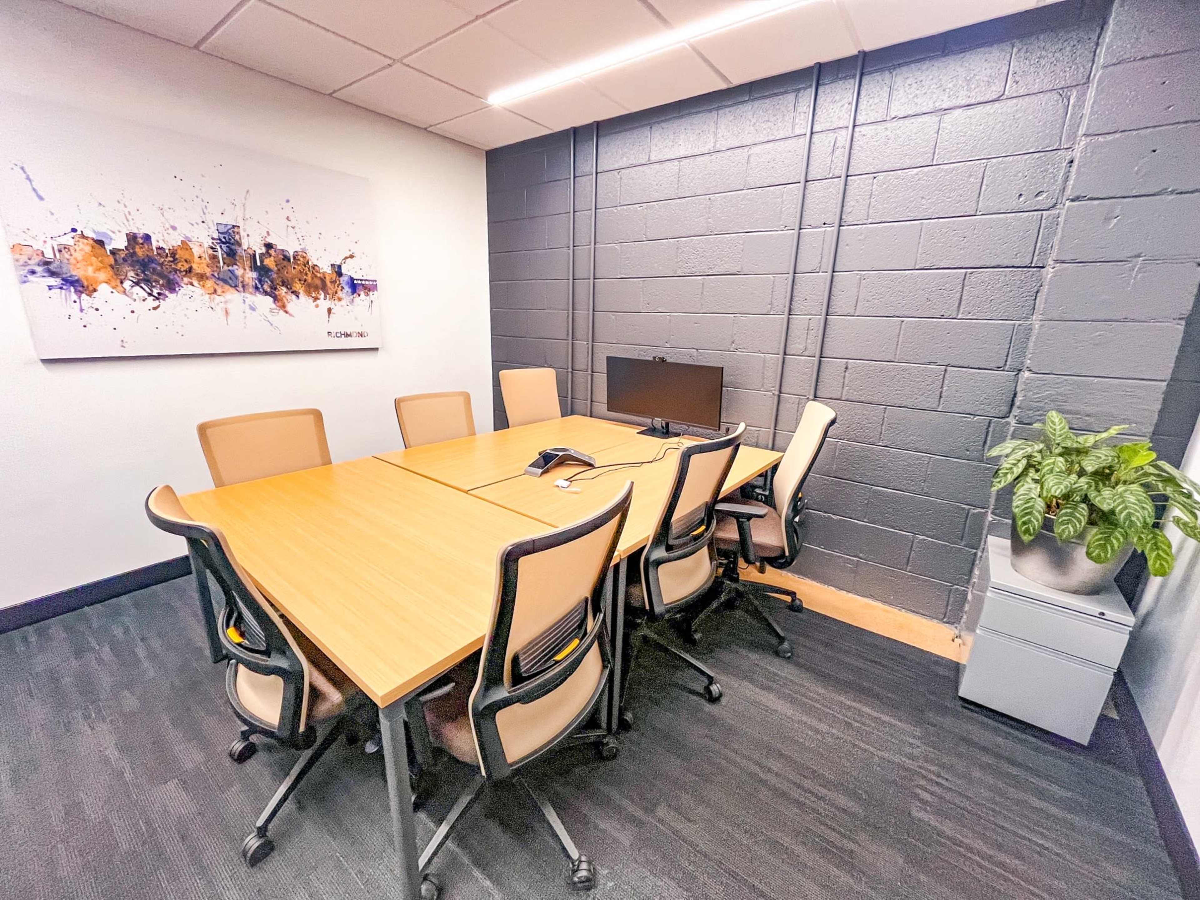 A conference room features a large wooden table surrounded by eight ergonomic chairs, with a monitor on one side and a potted plant in the corner.