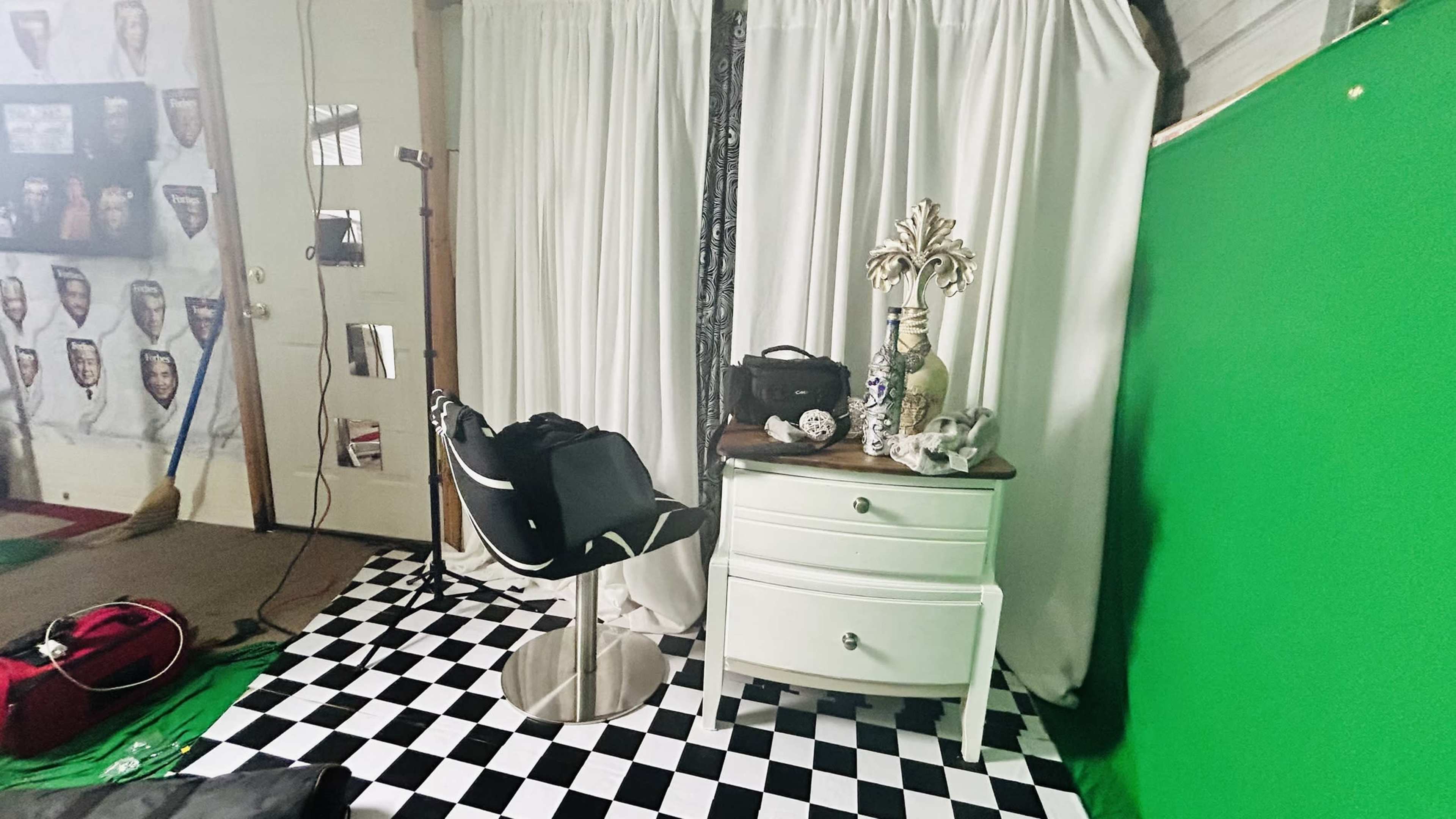 A small studio setup with a green screen, a black and white checkered floor, a vanity chair, a decorative table, and various objects arranged nearby.