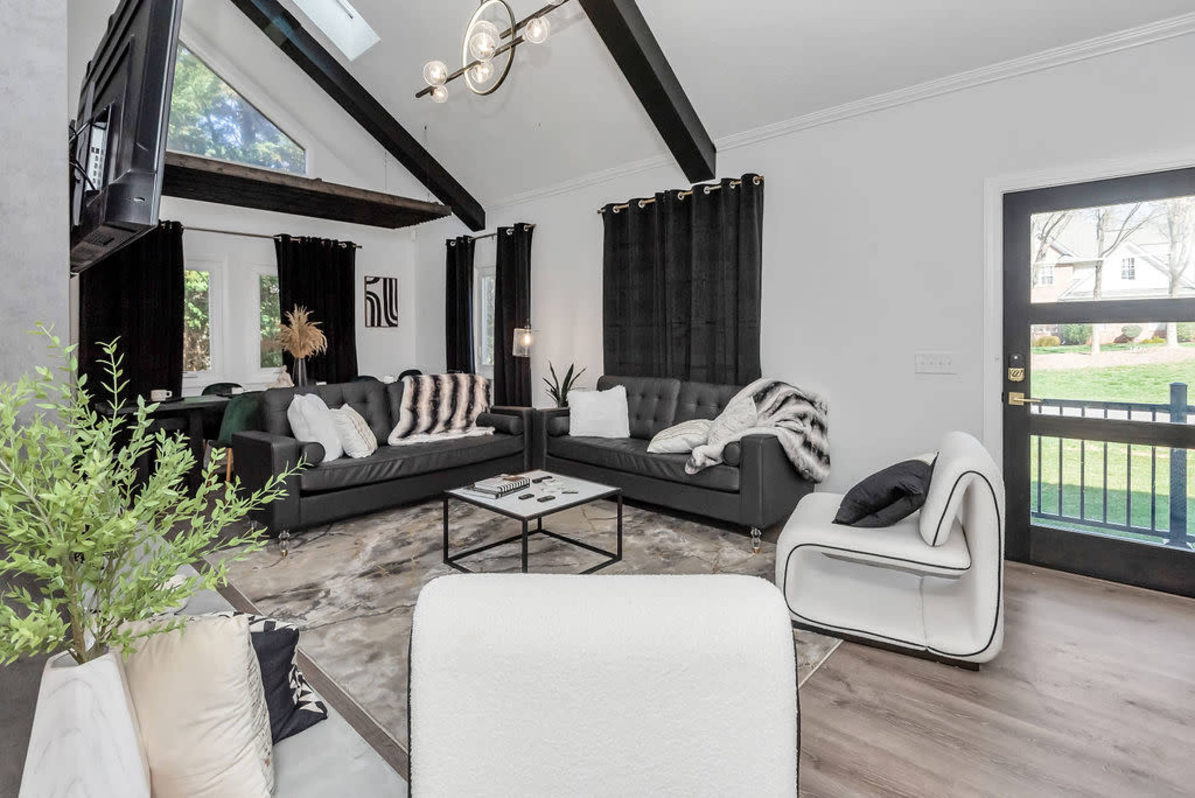 A modern living room features black sofas, a coffee table, and large windows with black curtains, all set beneath a vaulted ceiling with exposed beams.