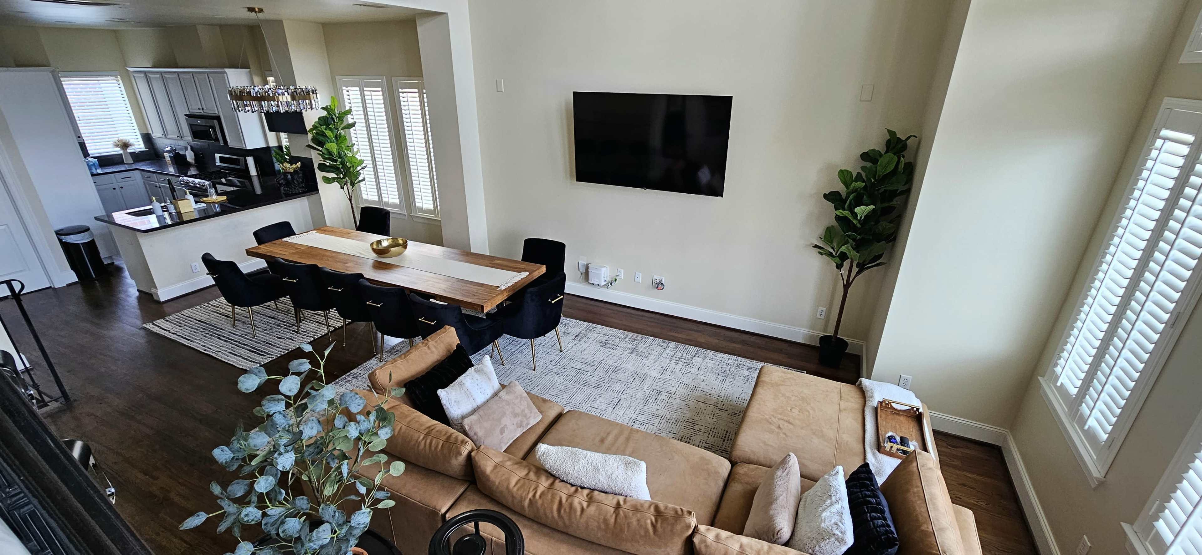 A spacious living area with a beige sofa, a wooden dining table surrounded by black chairs, and a large TV mounted on the wall.