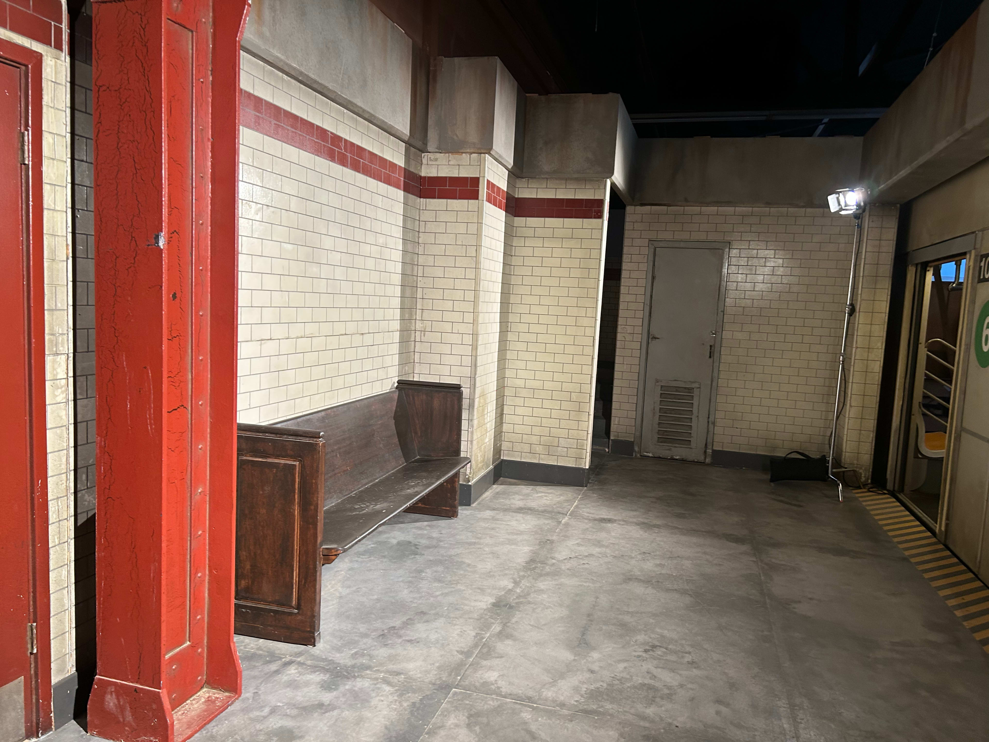 The image shows a subway station interior featuring a wooden bench against a white tiled wall, with red accents and a metal door in the background.