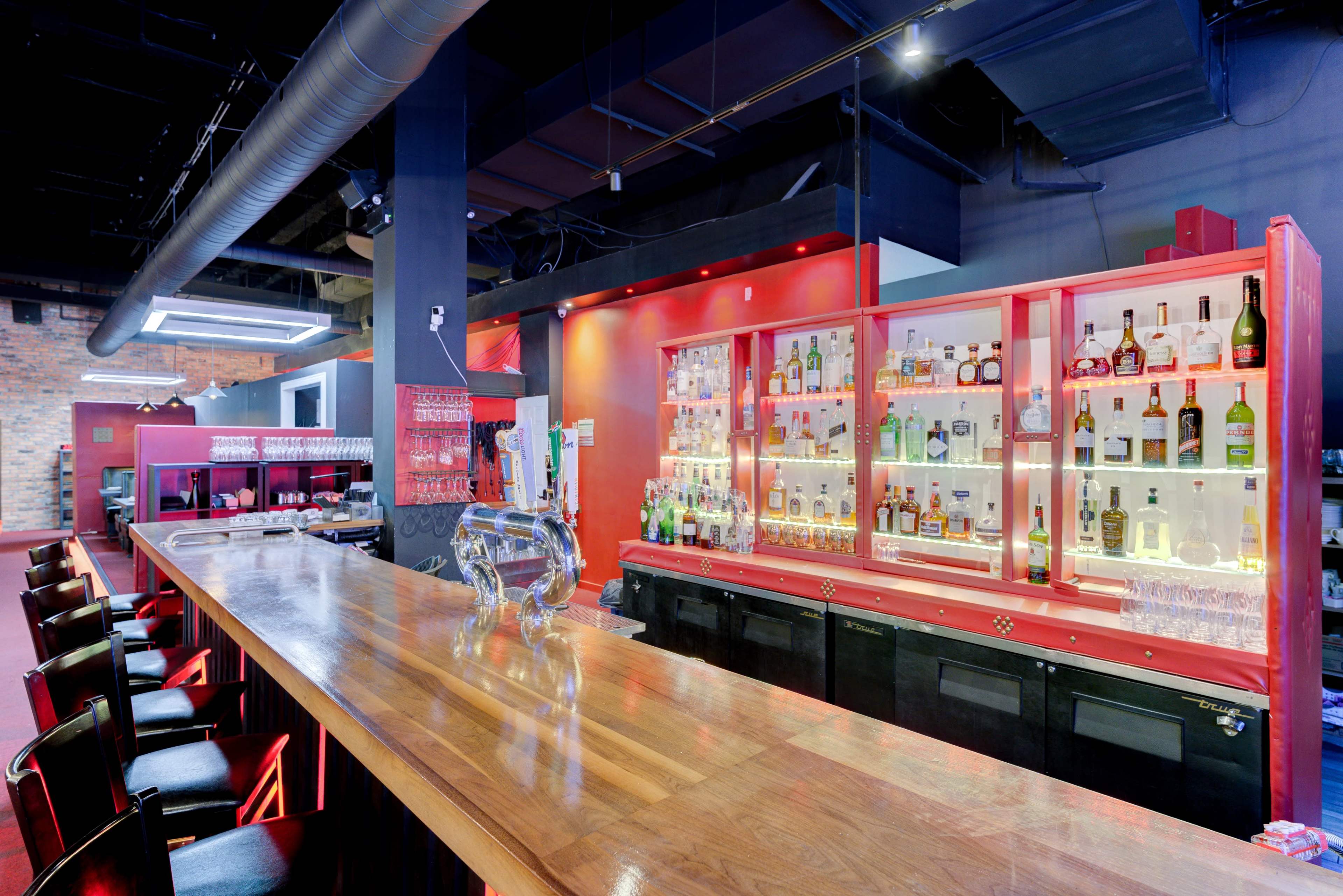 A modern bar with a wooden counter, glass shelves displaying various liquor bottles, and red accent lighting.