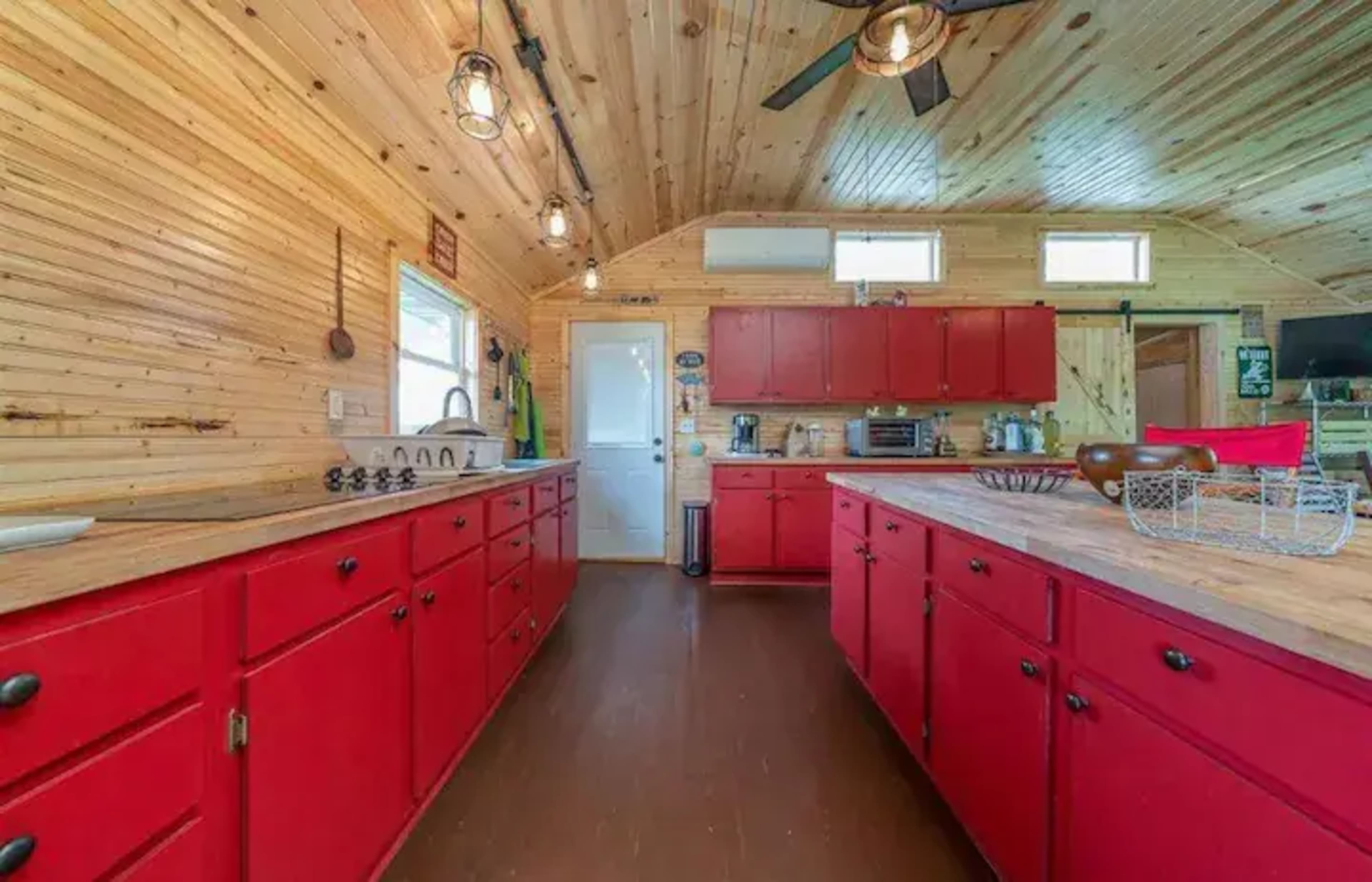 A spacious kitchen features red cabinets, wooden paneling, and large windows providing natural light.