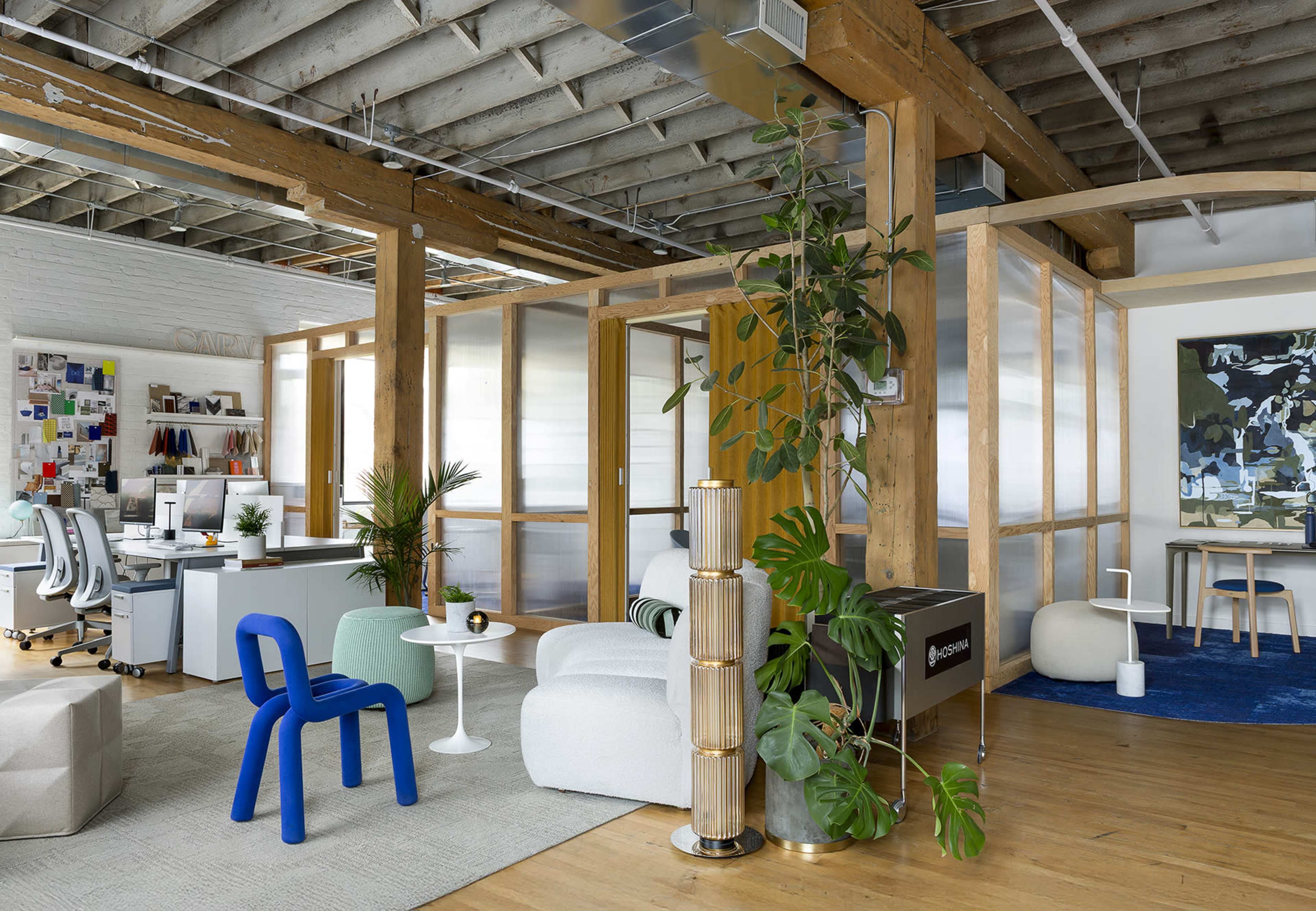 The image shows a modern office space featuring wooden beams, glass partitions, and various seating areas with plants and colorful furnishings.