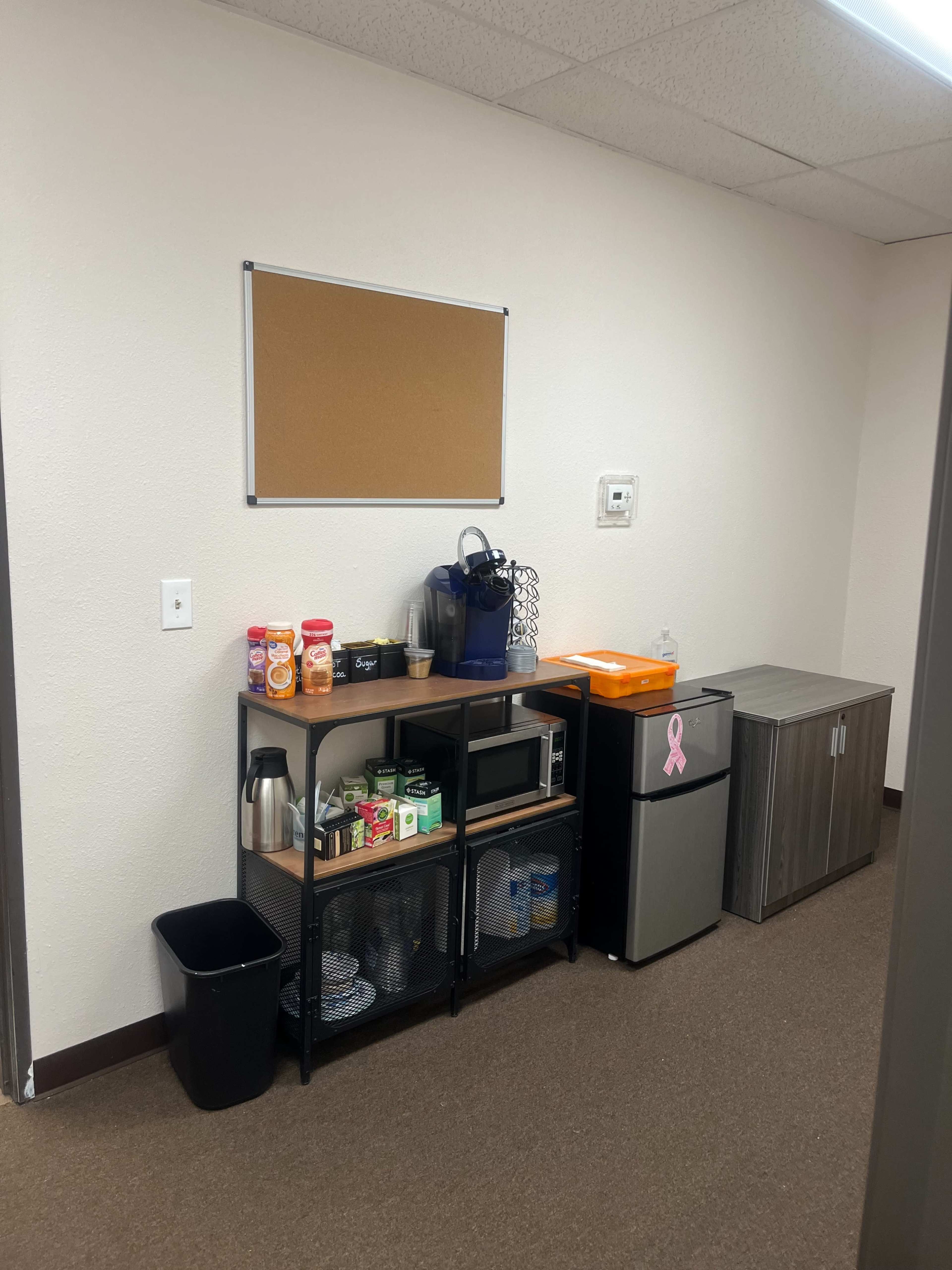 The image shows a small break area with a counter featuring a coffee maker, snacks, and a refrigerator, alongside a bulletin board on the wall.