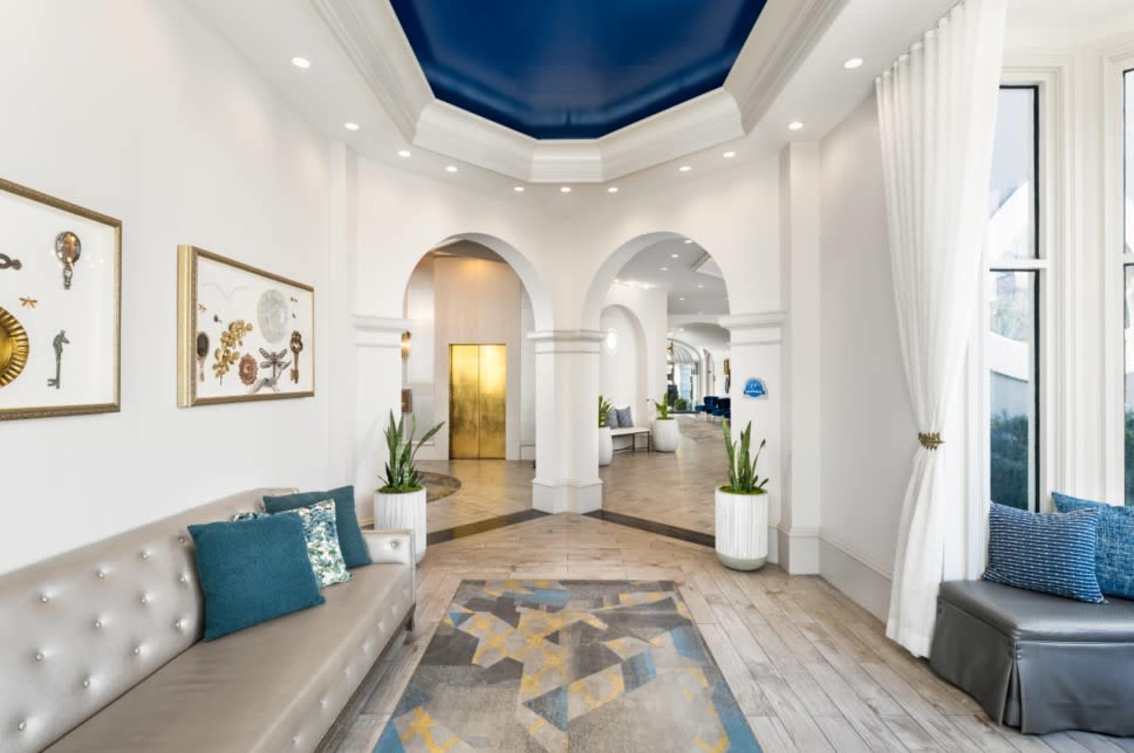 The image shows a spacious and well-lit lobby area with arched doorways, a decorative gray and yellow rug, and modern seating arrangements.