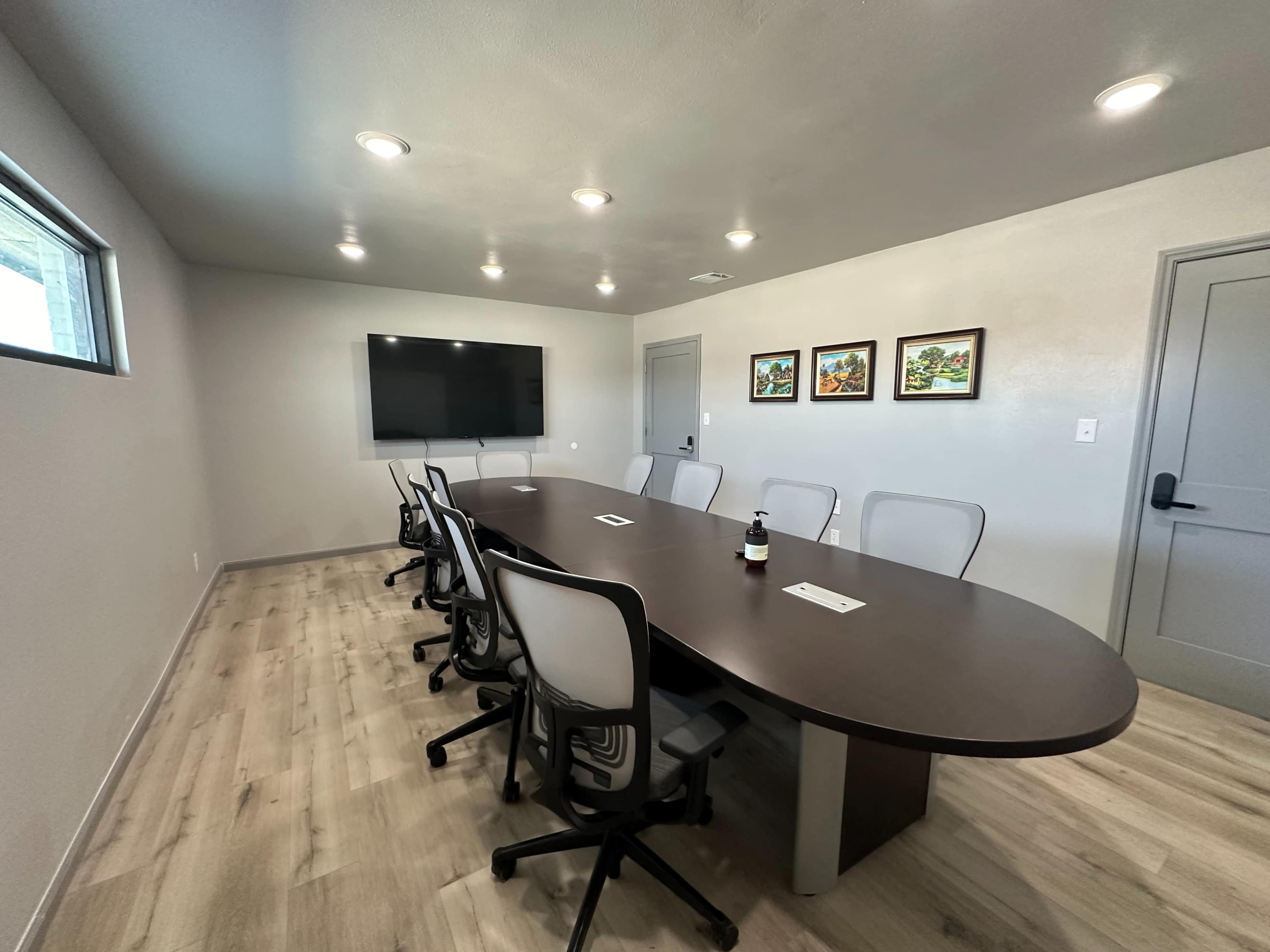 A sleek conference room features a large oval table surrounded by ergonomic chairs, with a wall-mounted television and framed artwork.