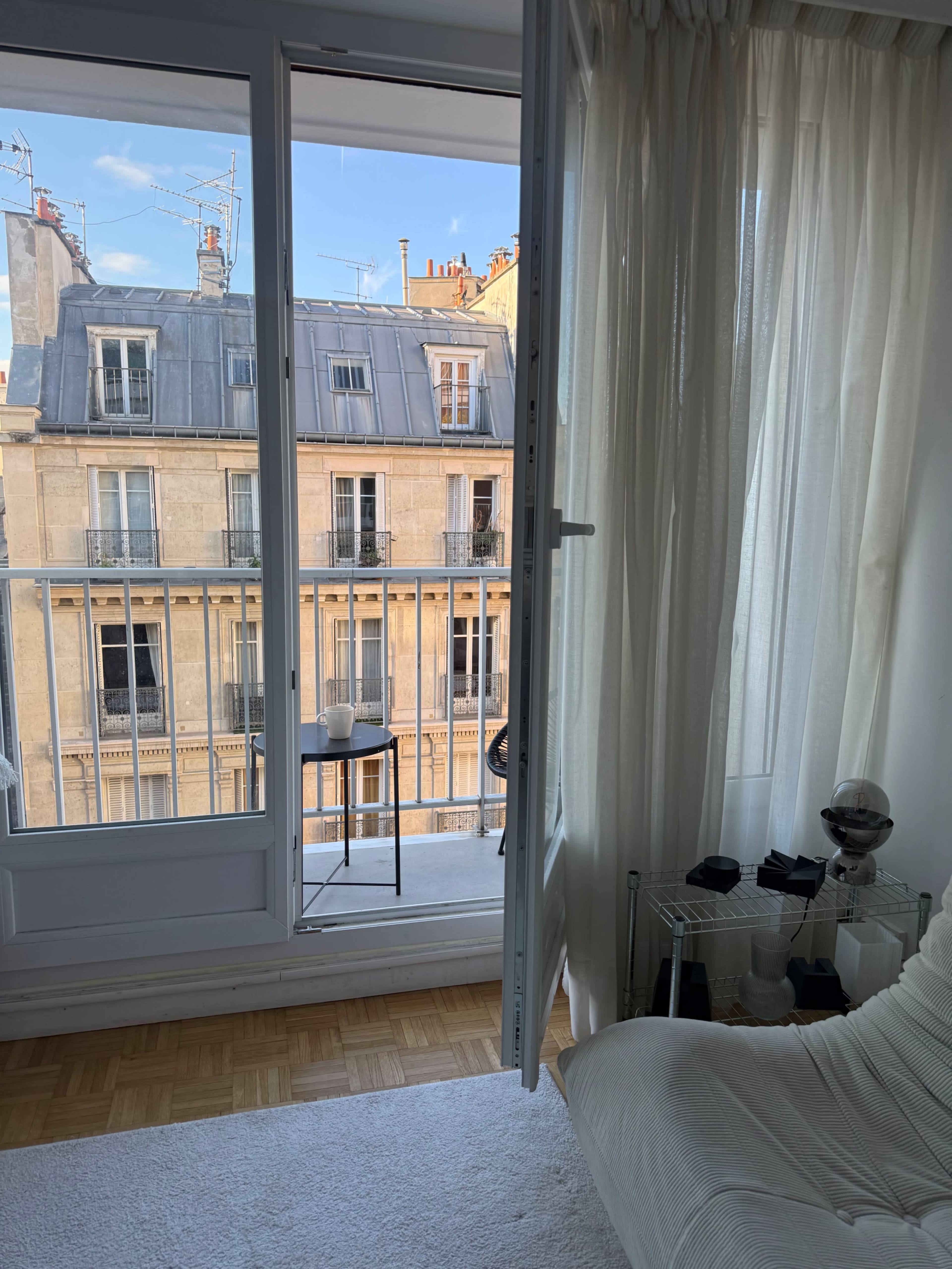 The image shows an open balcony door leading to a view of Parisian architecture, with a small table and chairs outside.