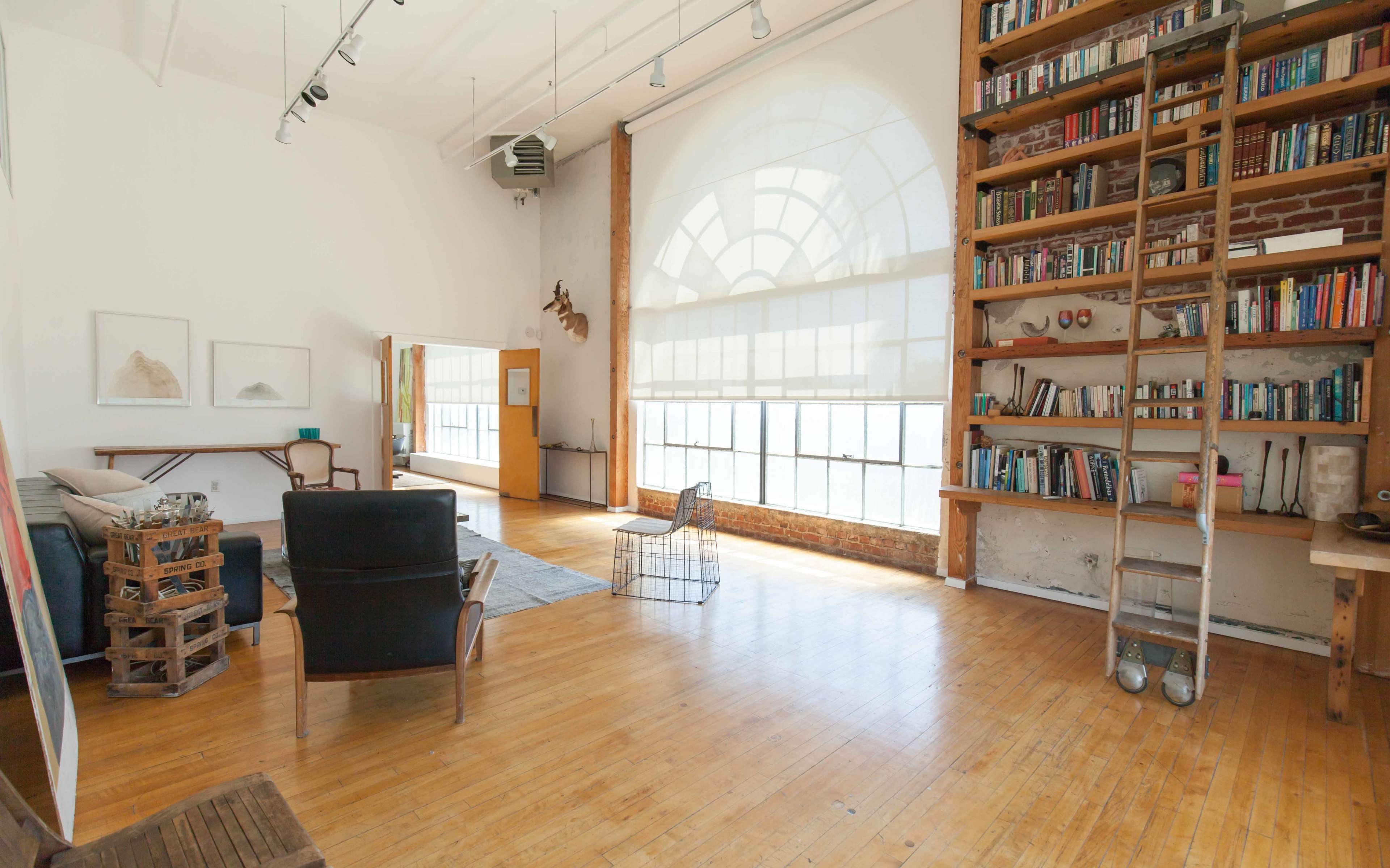 The image shows a spacious, brightly lit room with wooden flooring, featuring a large window, bookshelves, and minimalist furniture arrangements.