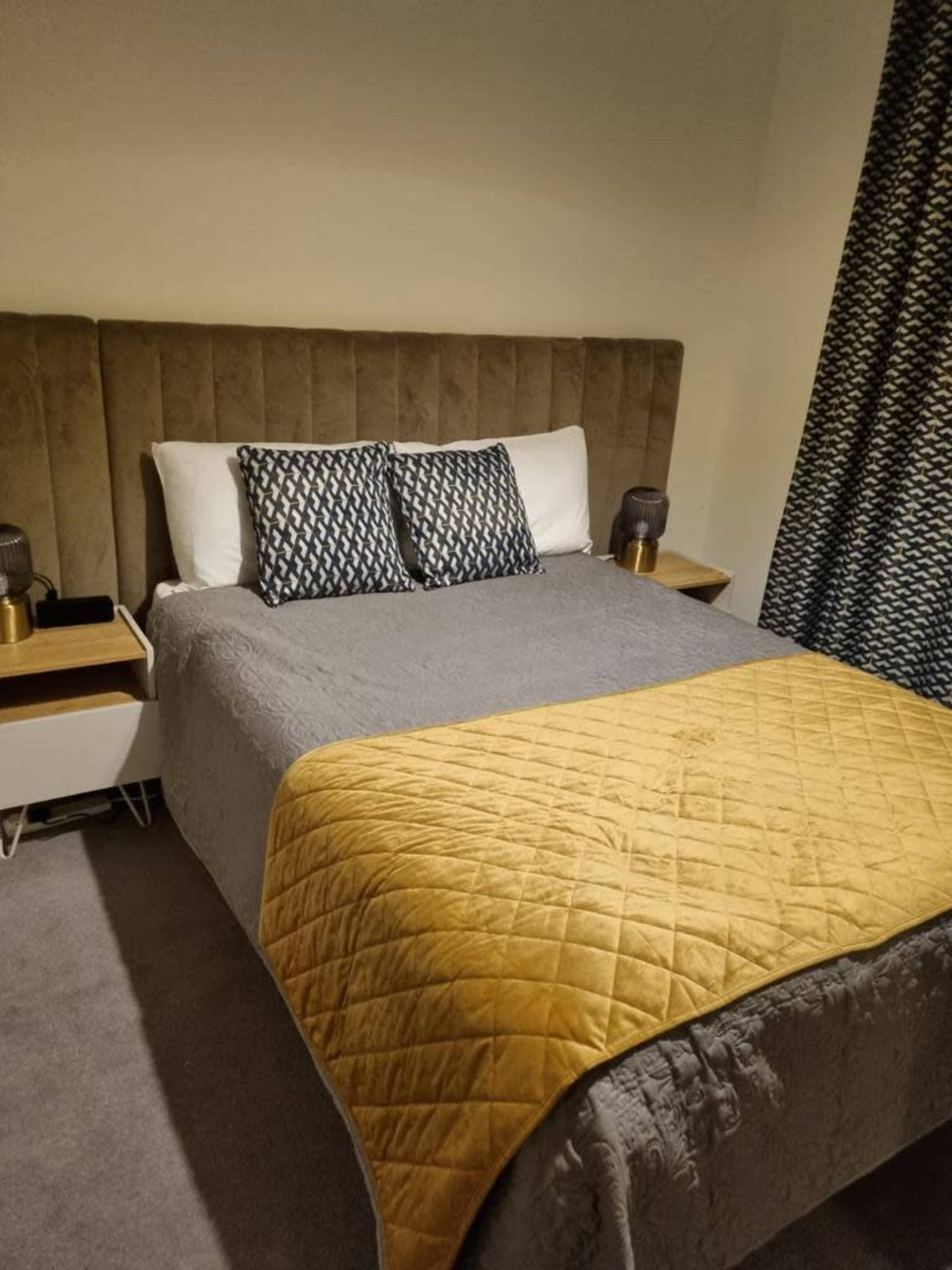 The image shows a neatly made bed with a gray quilt and a mustard-yellow throw, accompanied by two patterned pillows and bedside tables with lamps.