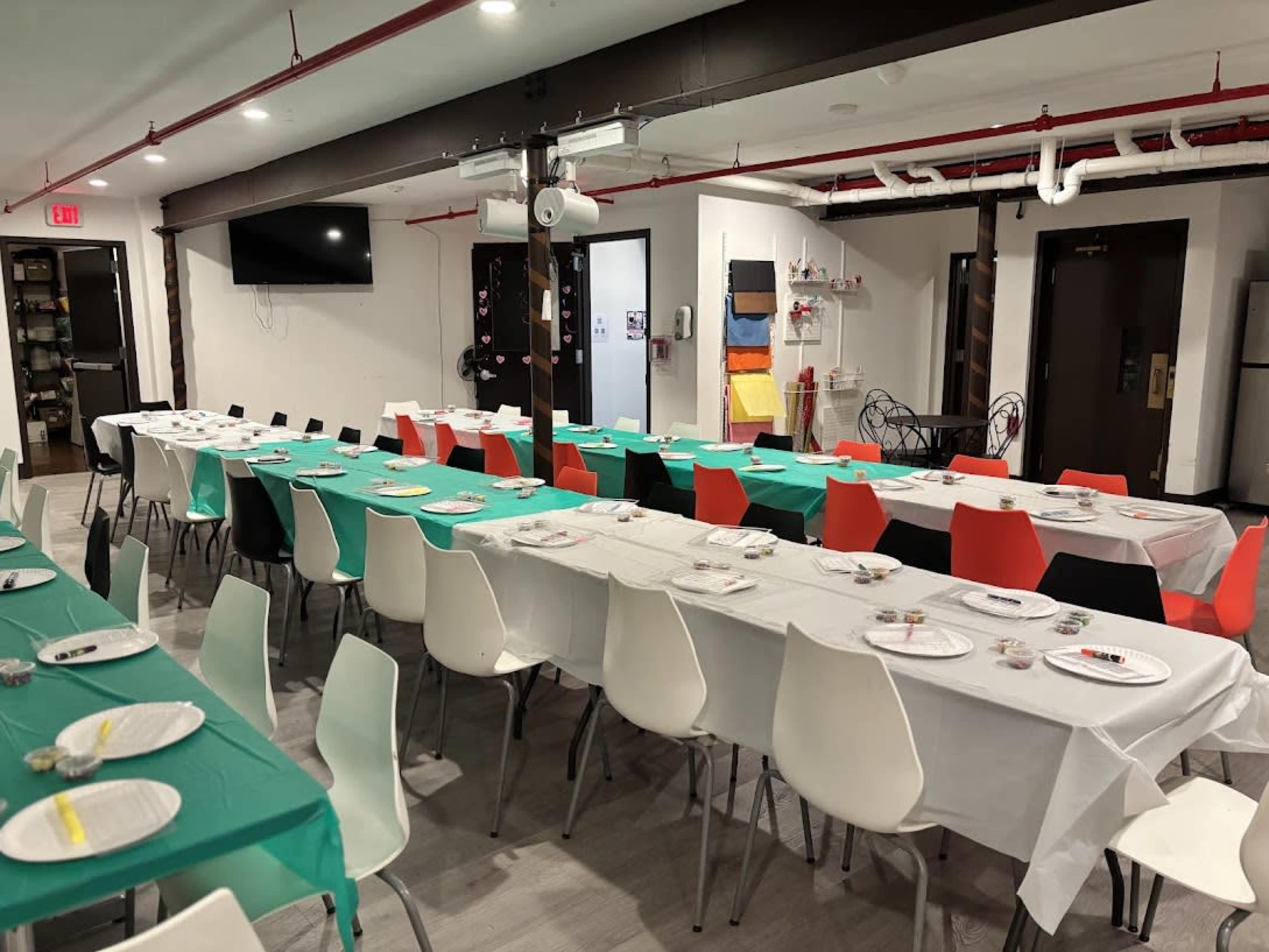A long dining table is set with white and green tablecloths, decorated with plates and colorful chairs arranged in a spacious room.