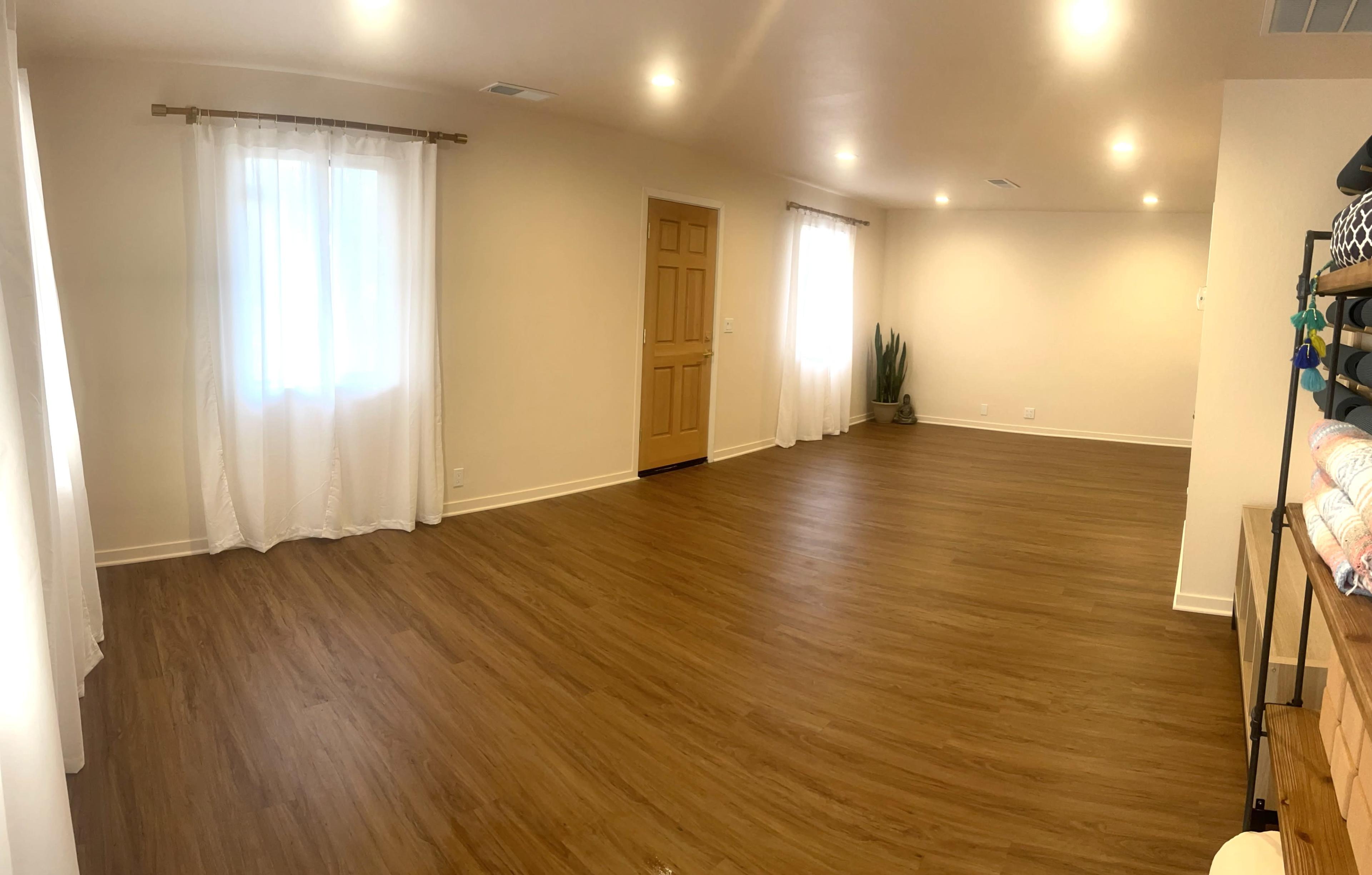 A spacious room with light-colored walls, wooden flooring, a large window with sheer curtains, and minimal furnishings.