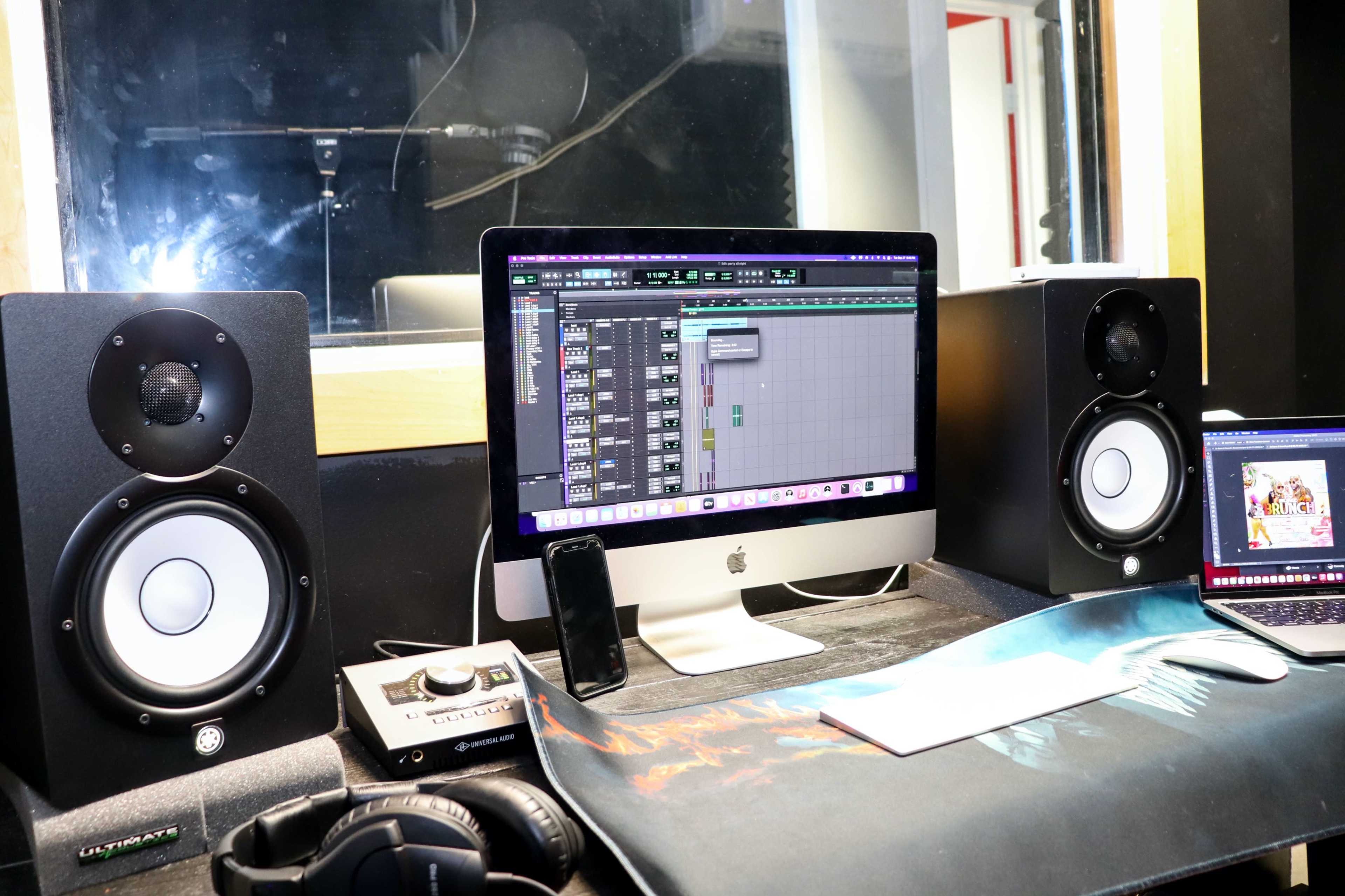The image shows a music production workstation featuring a desktop computer with audio software open, two speakers, a mixing interface, headphones, and a laptop.