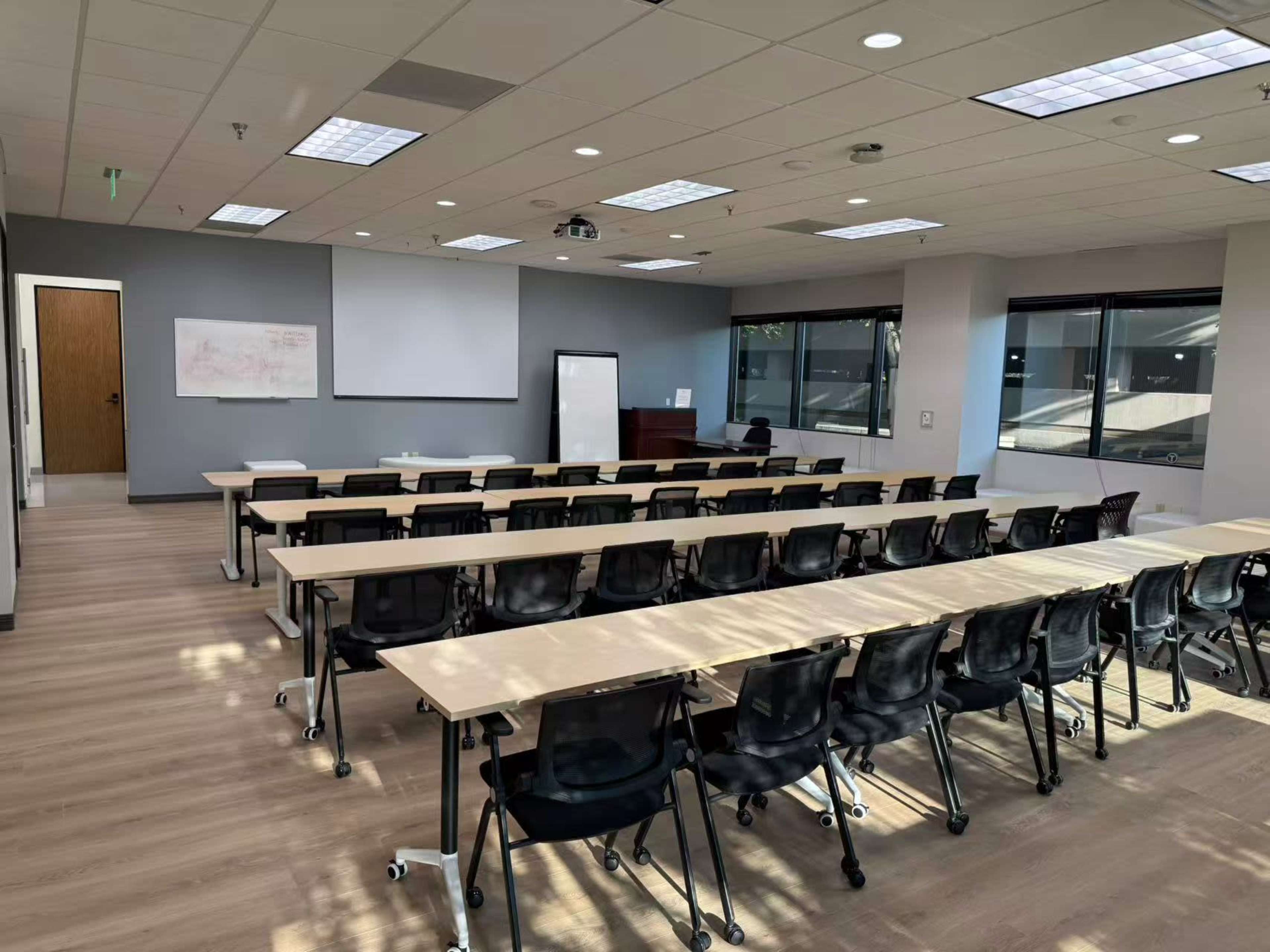 The image shows a modern classroom with several rows of tables and chairs, a whiteboard, and a projector at the front.
