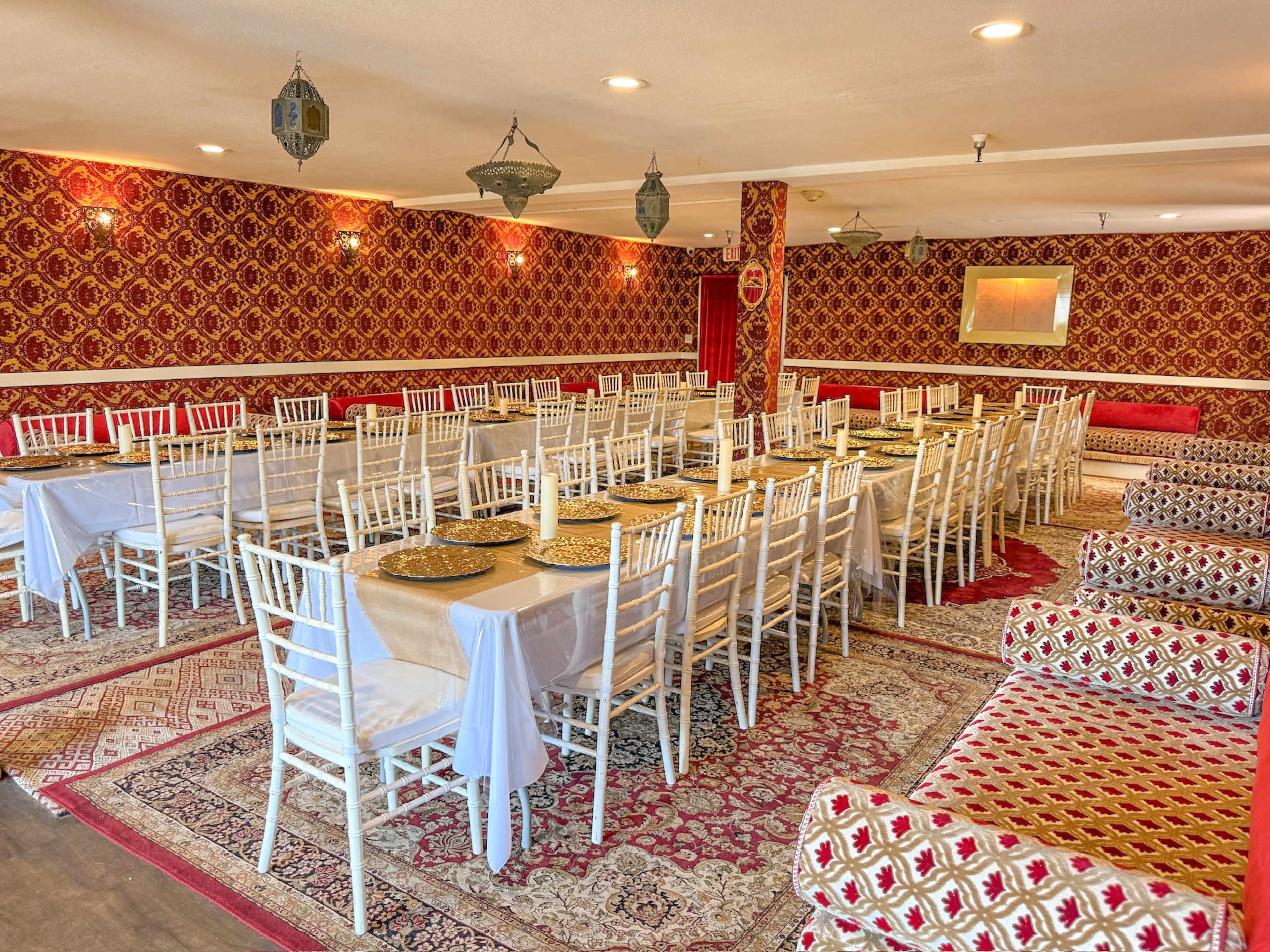 The image shows a spacious dining area with rows of white-topped tables set with gold chargers, surrounded by ornate red patterned walls and decorative lighting.
