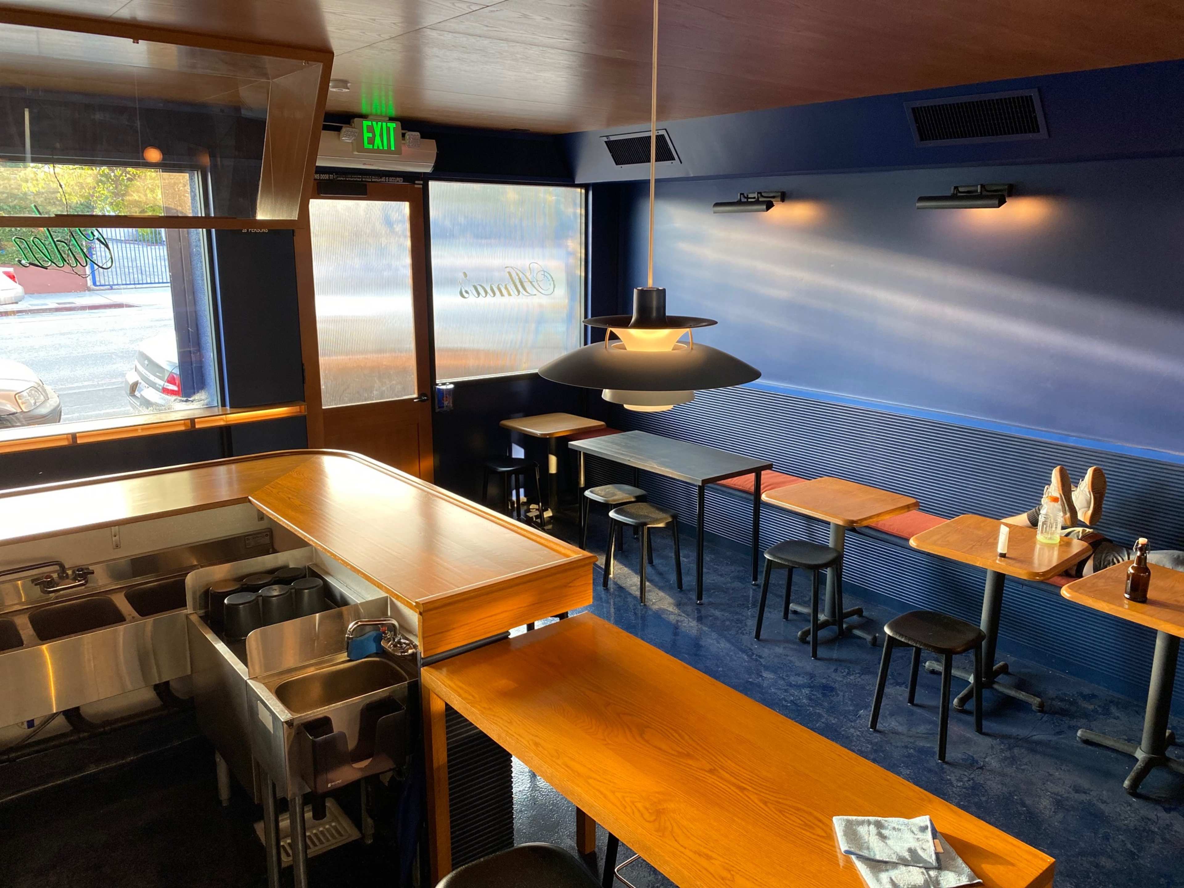 The interior of a modern restaurant with a wooden counter, black stools, and tables against a blue wall.