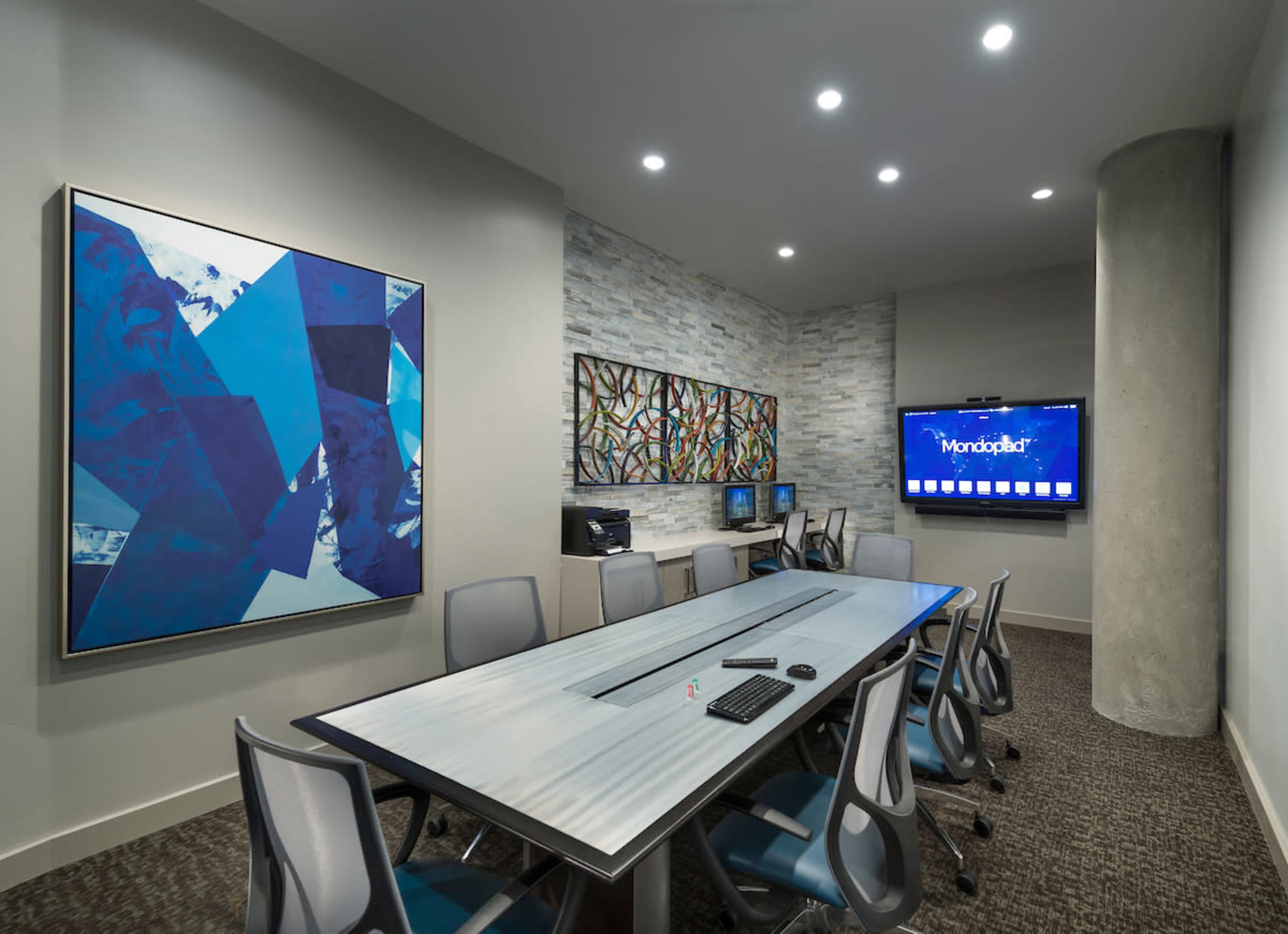 A modern conference room with a large table, several chairs, and two wall-mounted artworks, along with a television and computers in the background.