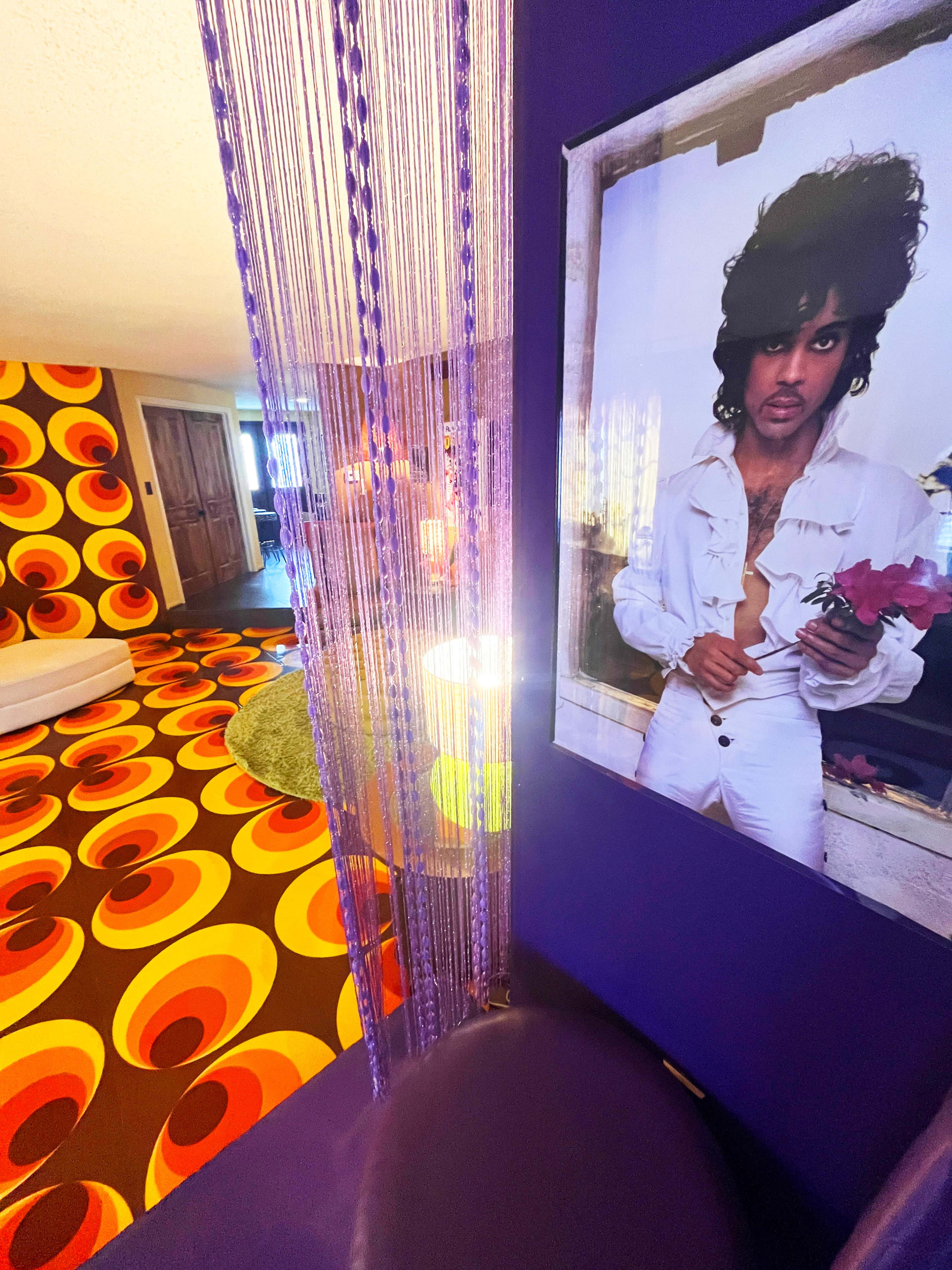 The image shows a vibrant room featuring a bold, retro-patterned carpet and a prominent portrait of a musician against a purple wall.