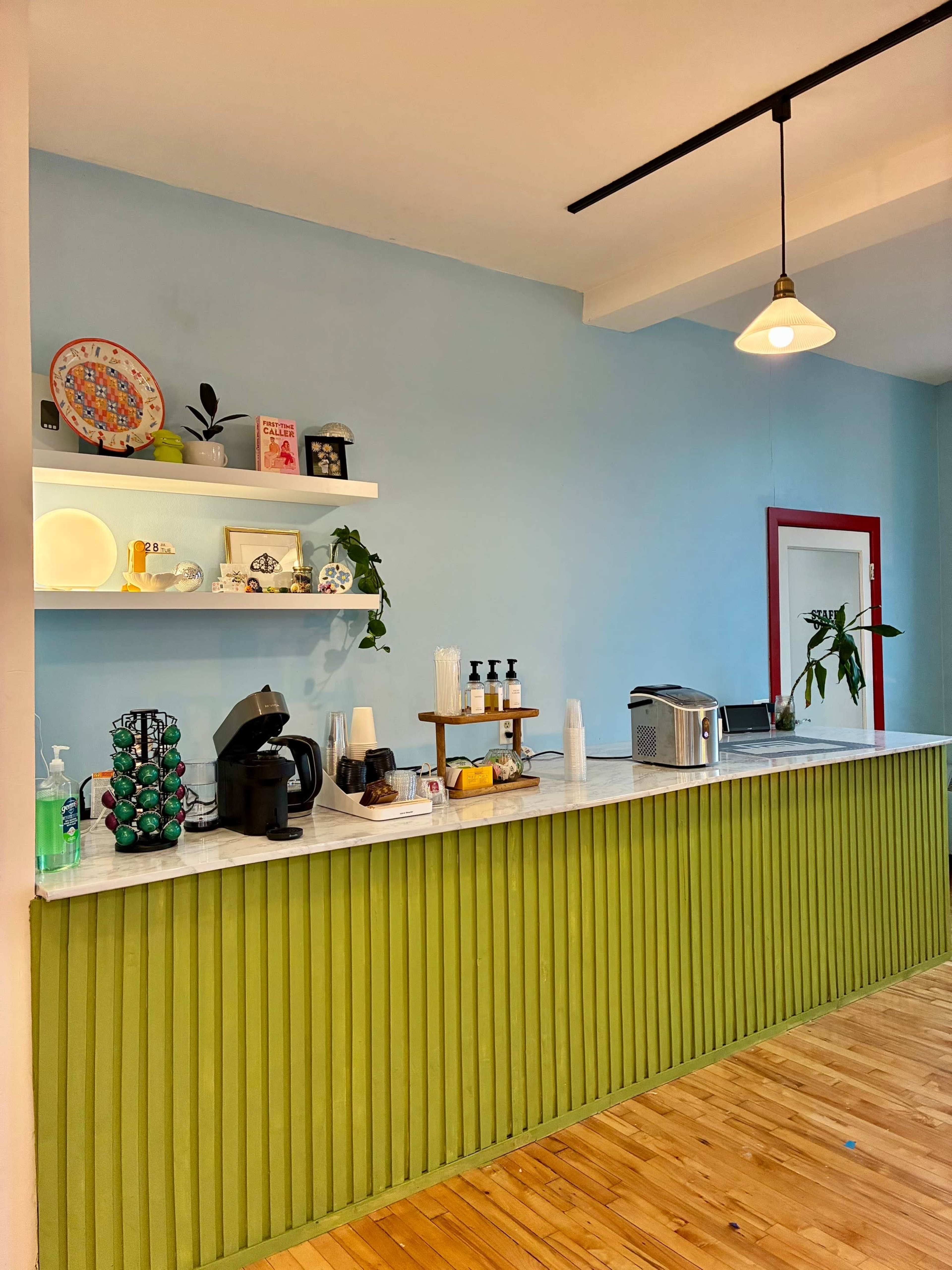 The image shows a café counter with a green panelled front, a coffee maker, and decorative items on shelves against a light blue wall.