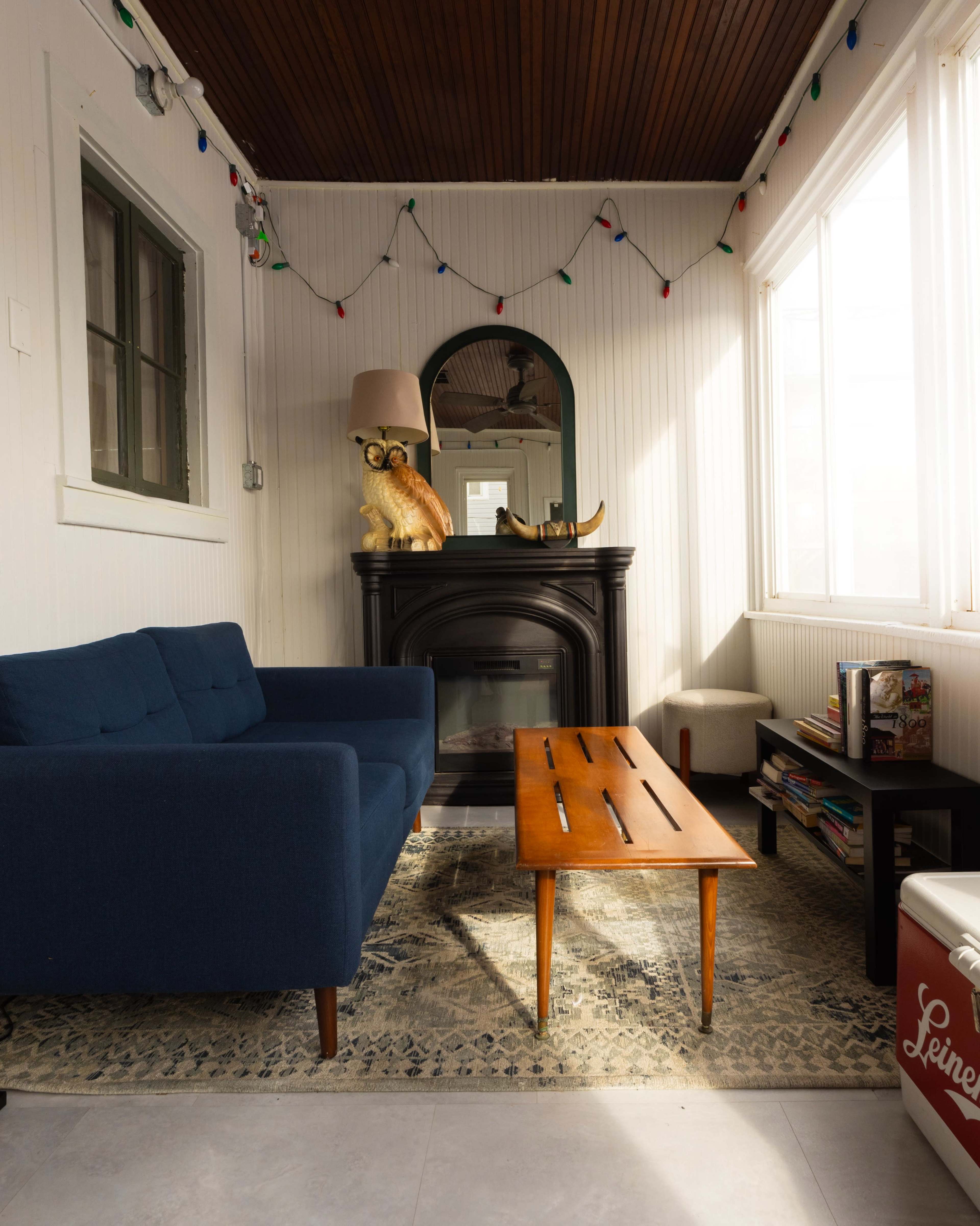 The image shows a cozy living area with a blue sofa, a wooden coffee table, and a decorative fireplace, all featuring warm lighting and a few colorful string lights overhead.
