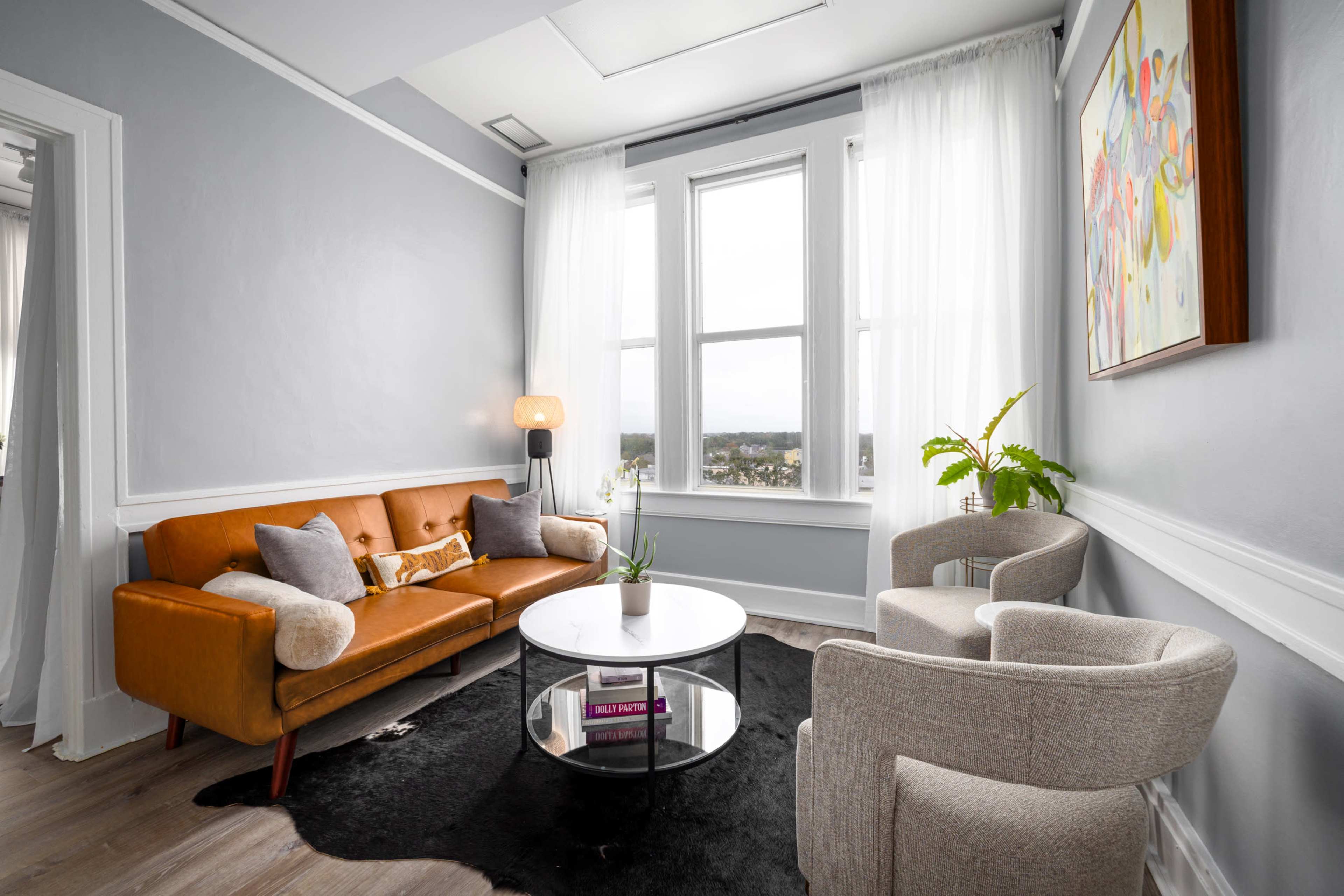 The image shows a modern living room with a brown leather sofa, a circular glass coffee table, two light gray armchairs, and large windows with sheer white curtains.