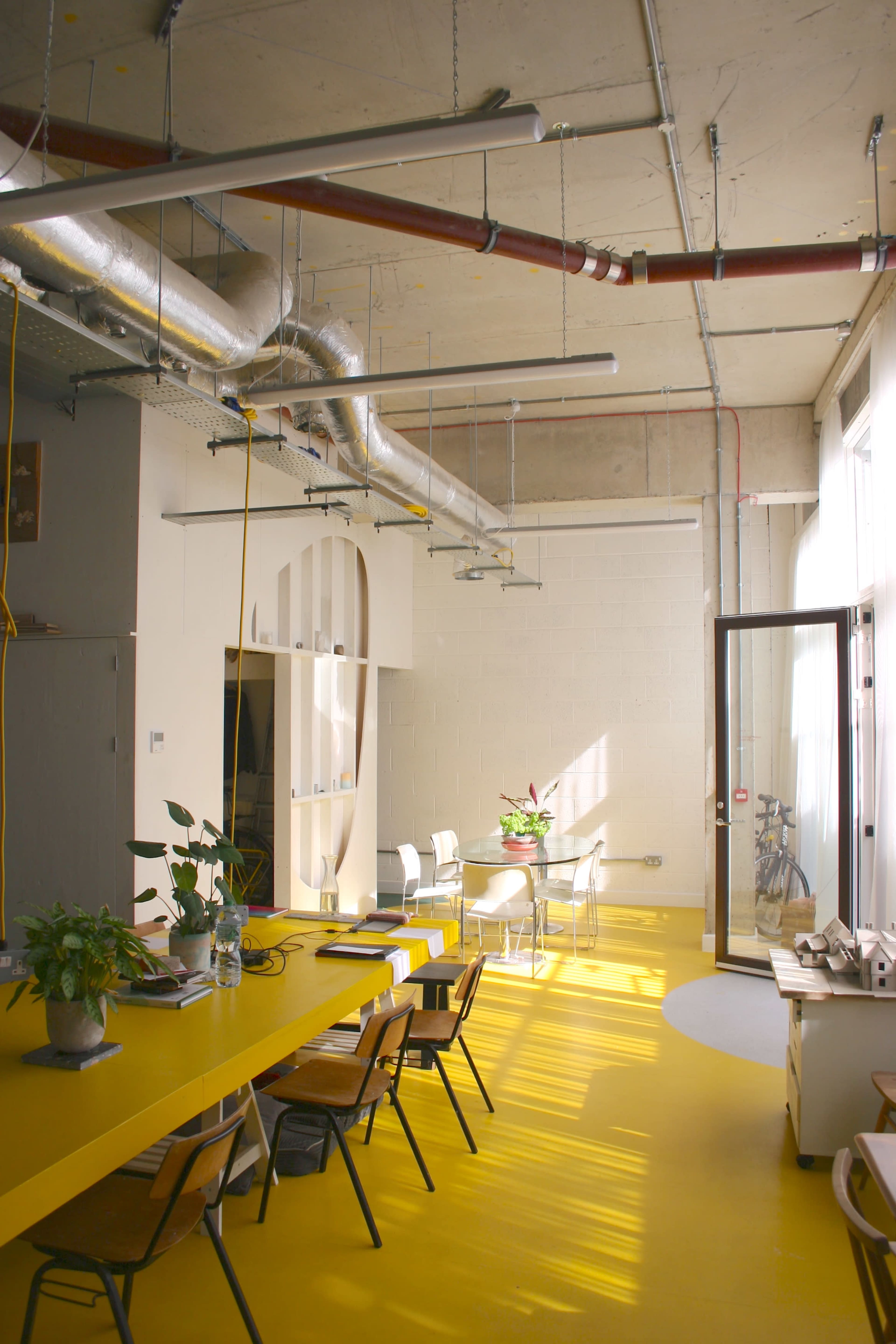 The image shows a bright, modern office space with a long yellow table, several chairs, and large windows letting in natural light.