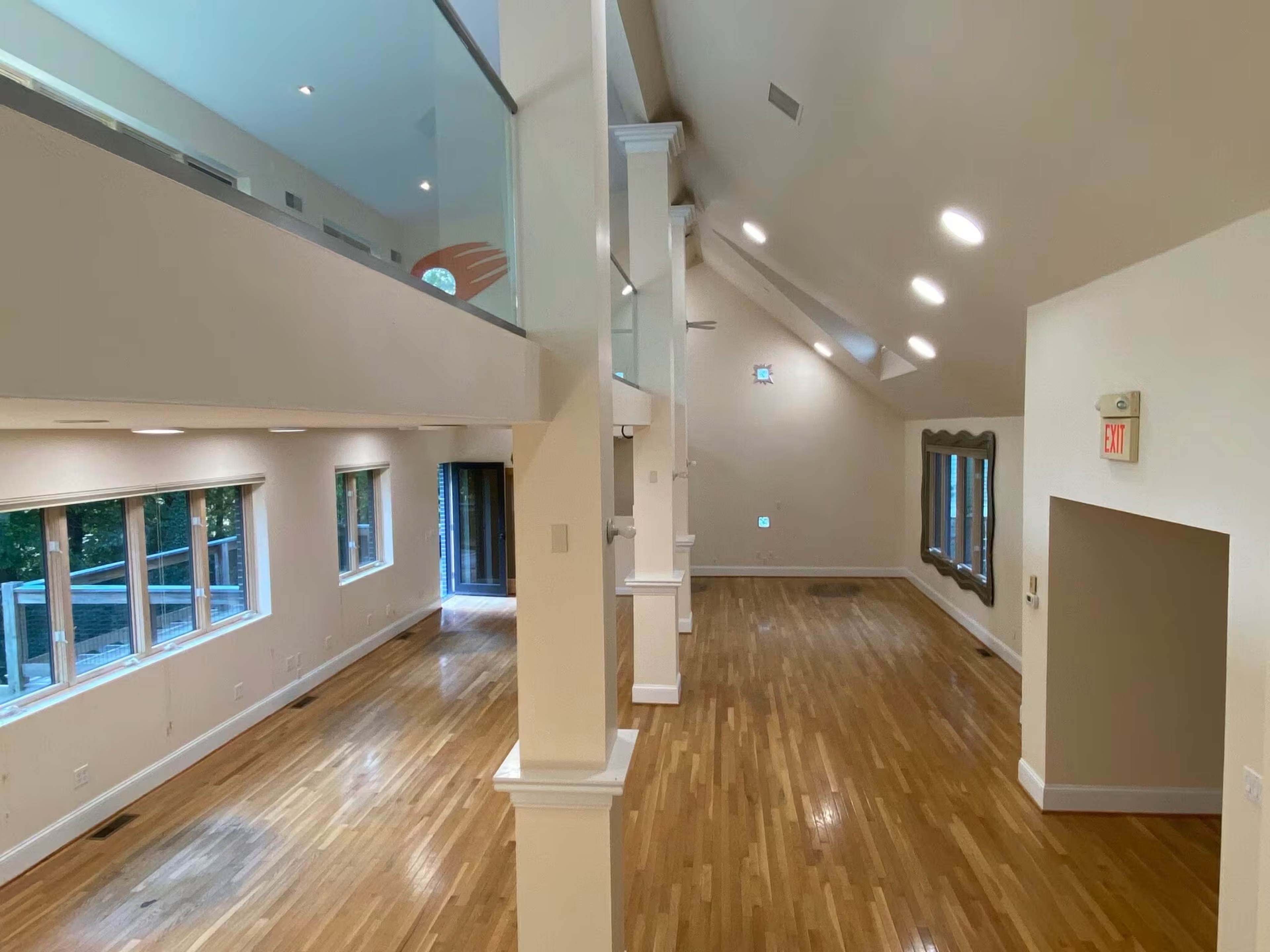 The image shows a spacious, empty room with wooden floors, tall ceilings, and large windows allowing natural light to enter.