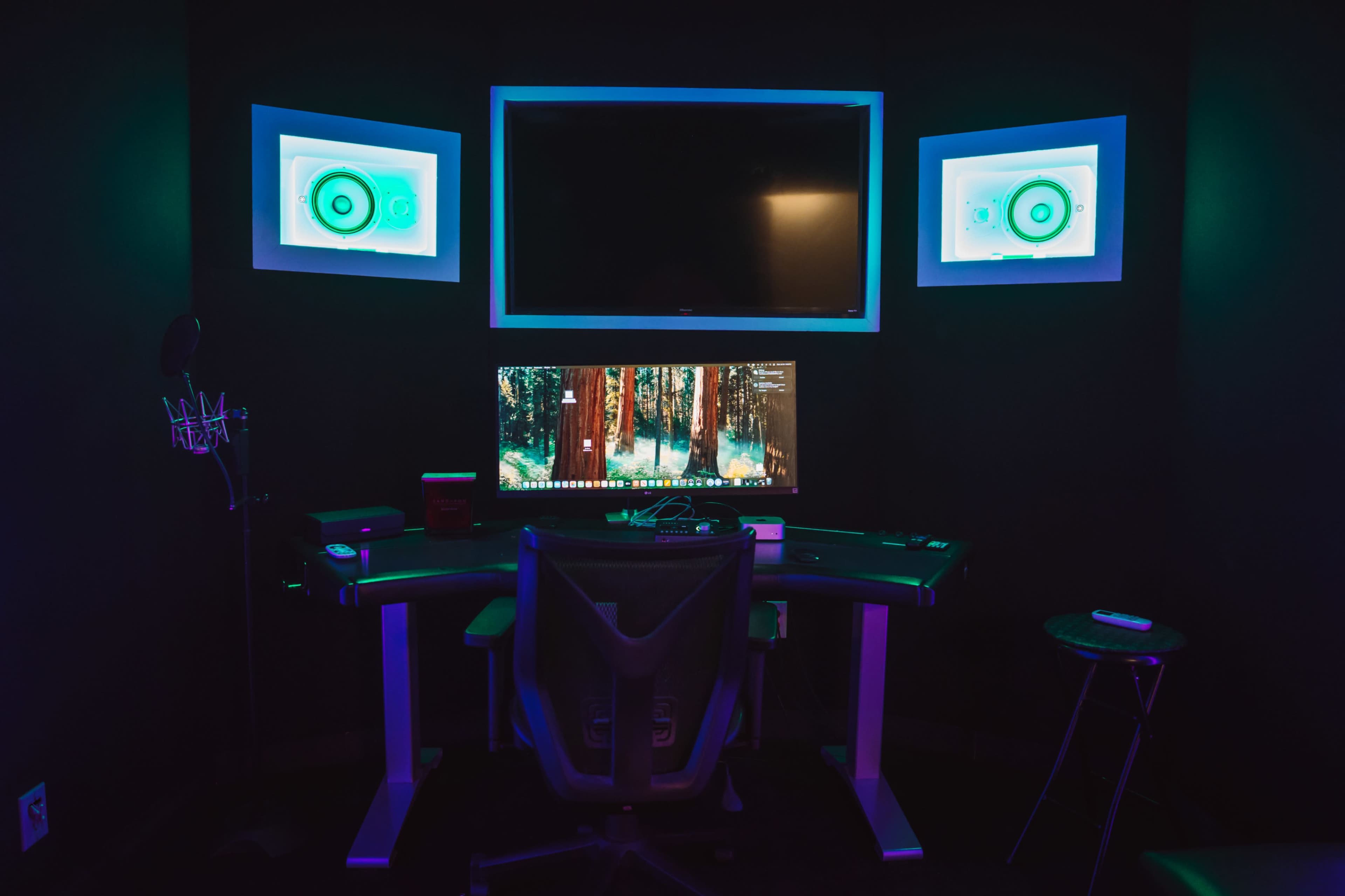 The image shows a dark room with a desk, computer monitor, and a large screen mounted on the wall, illuminated by blue and green lights.