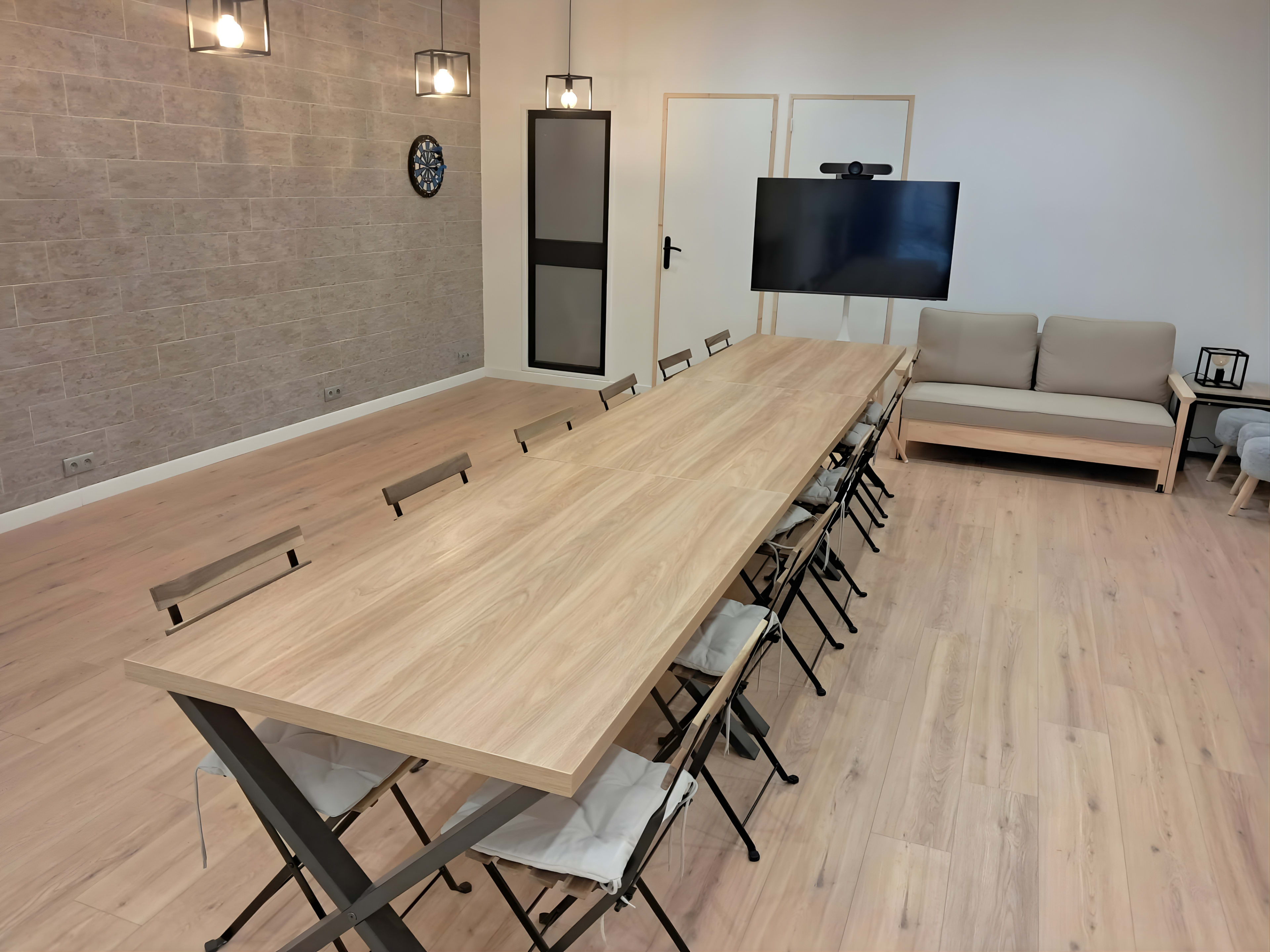 A long wooden table with metal legs is set in a well-lit meeting room, surrounded by several chairs, with a sofa and a television on one side of the room.