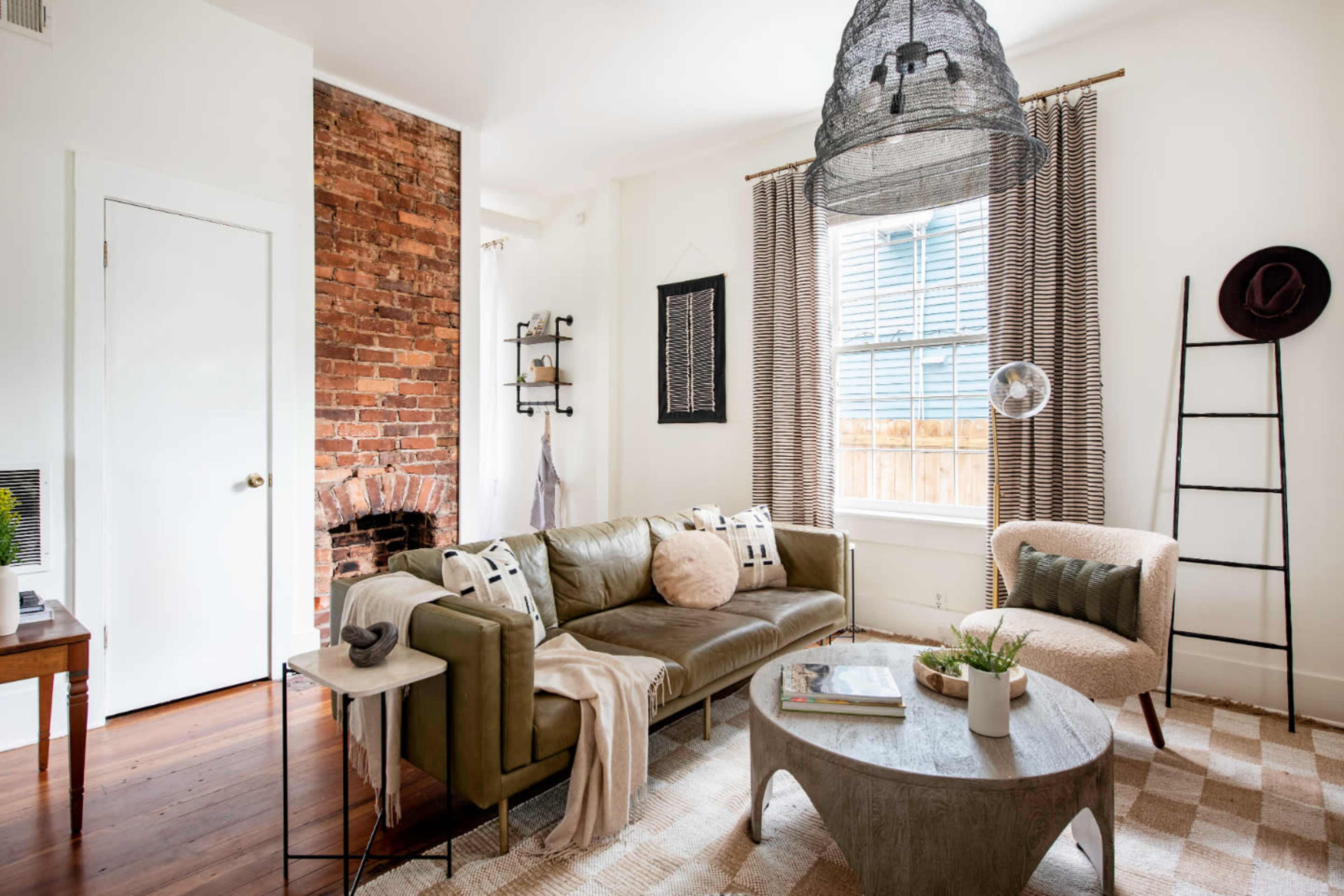 The image shows a cozy living room featuring a green leather sofa, a light-colored armchair, a round coffee table, and a brick accent wall with large windows.