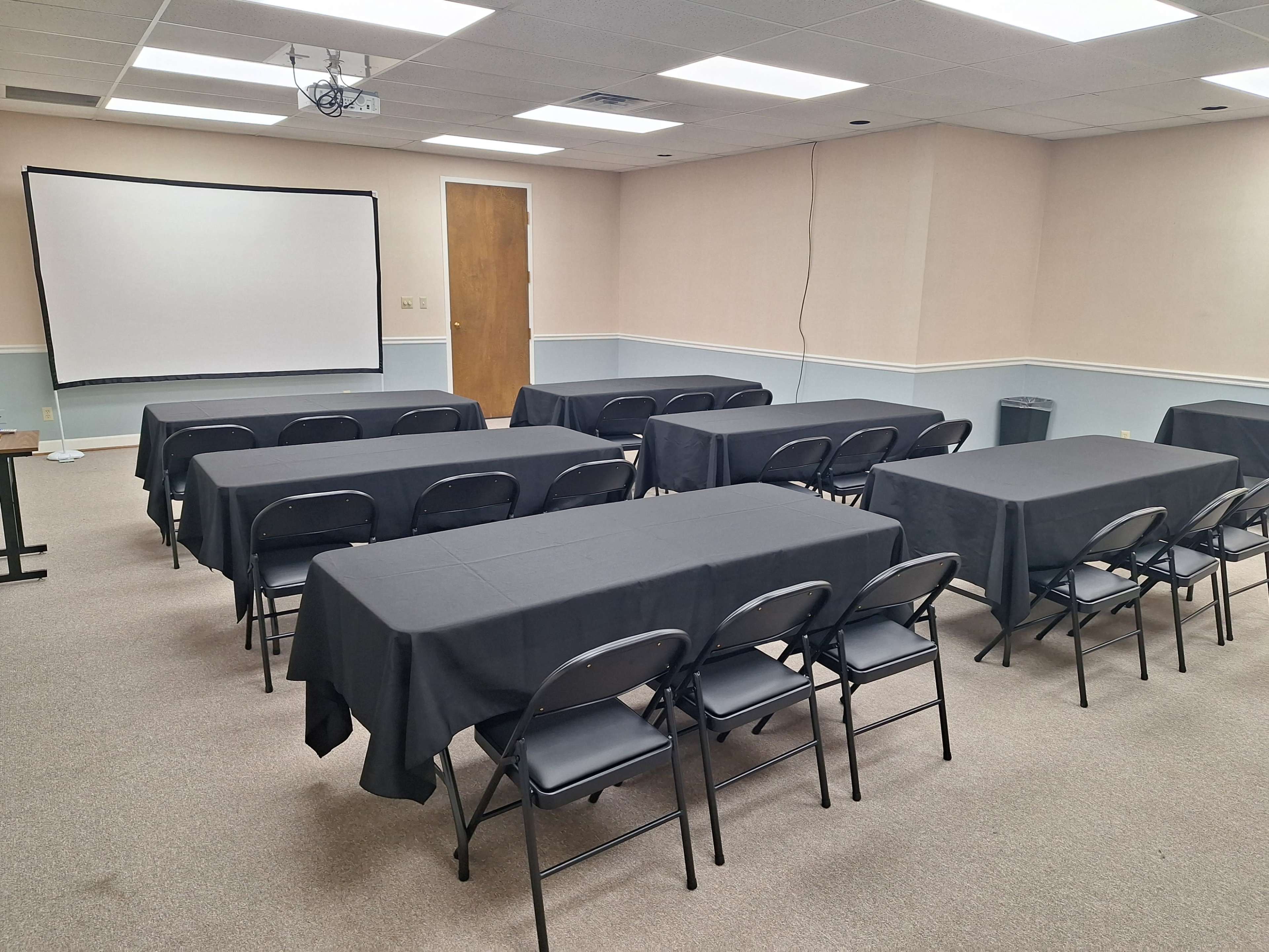 A meeting room is set up with several rows of tables covered in black tablecloths and chairs arranged around them, along with a projector screen at the front.