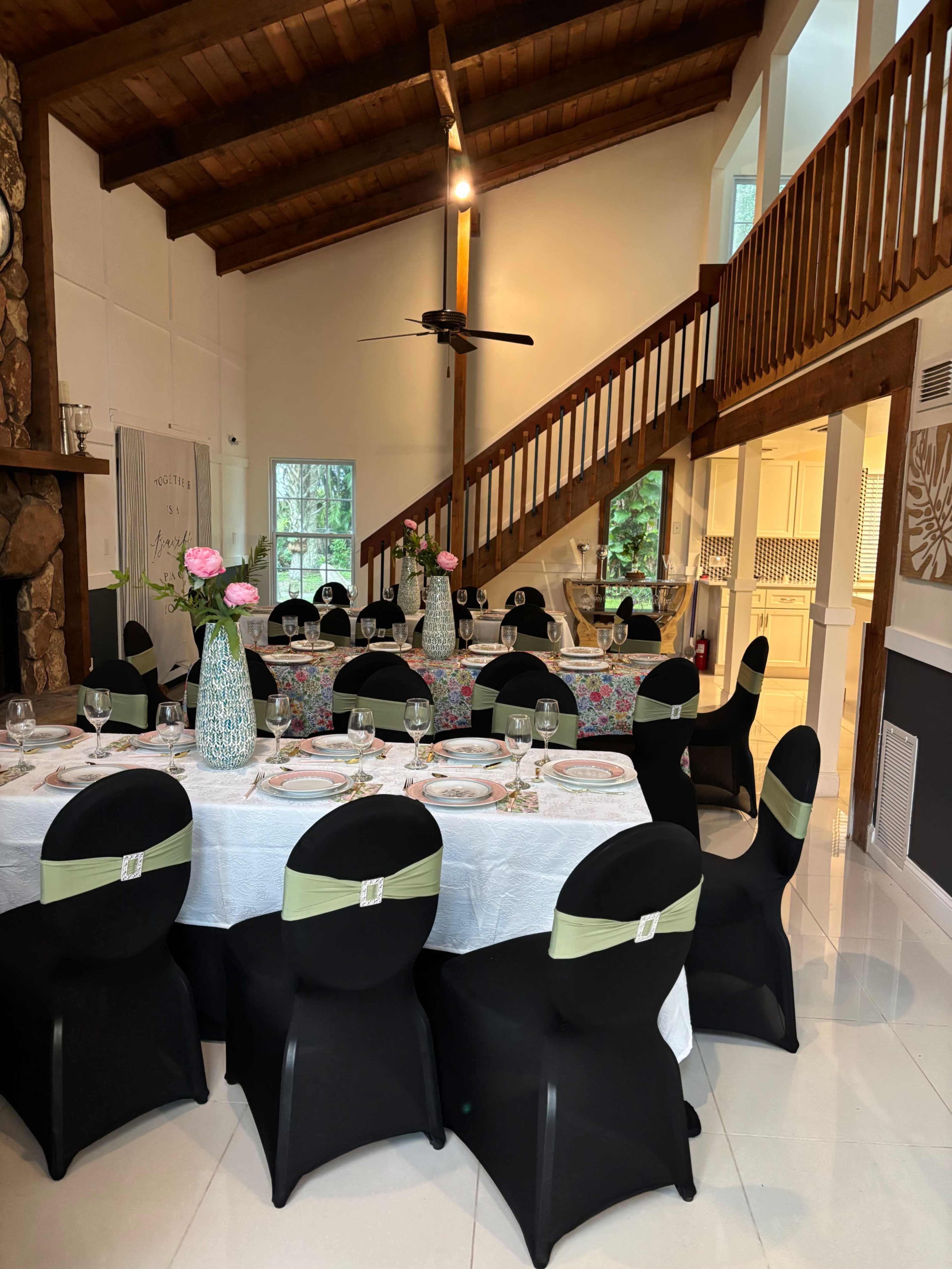 A dining area features several round tables covered in white tablecloths with black chairs adorned with green sashes, illuminated by natural light from nearby windows.