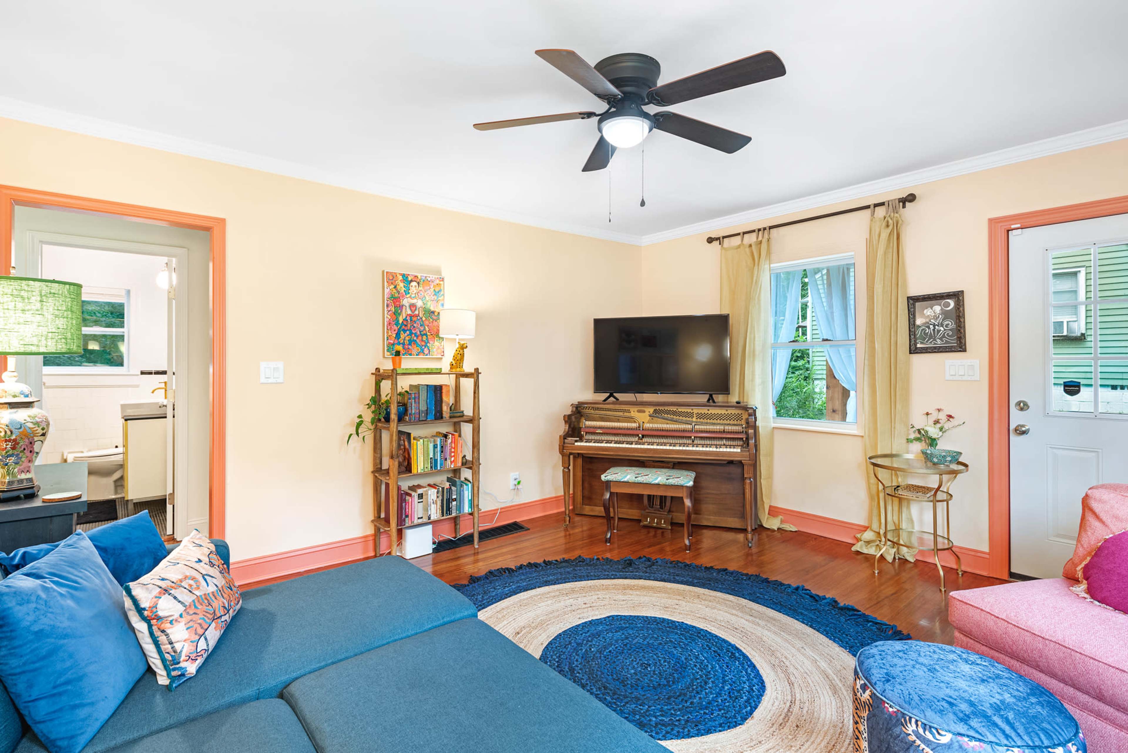 The living room features a blue sectional sofa, a wooden piano, a television, and a circular area rug, surrounded by light-colored walls and colorful accents.