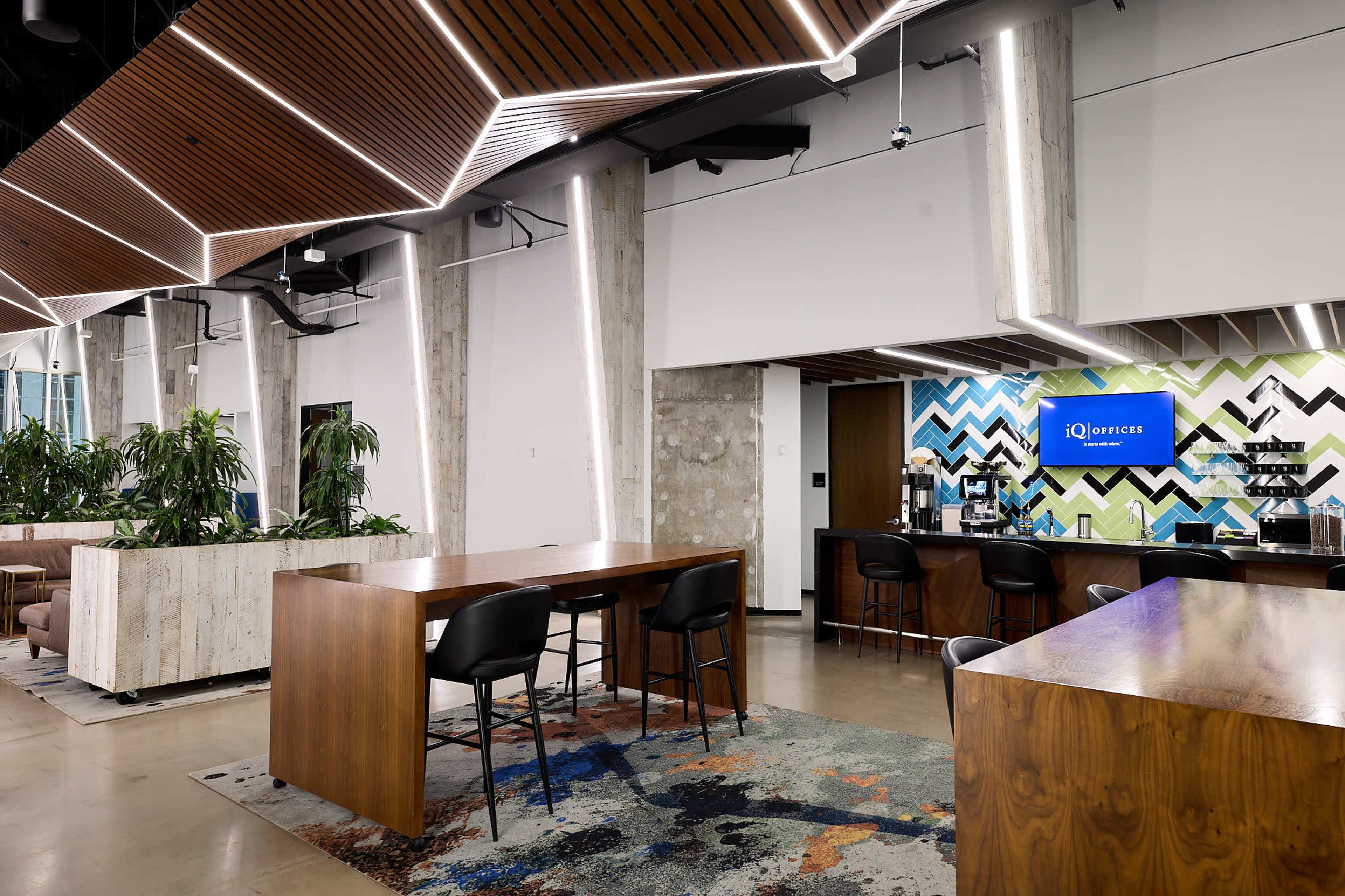 The image shows a modern office space featuring wooden tables, a coffee bar, and decorative plants against a backdrop of geometric wall patterns.