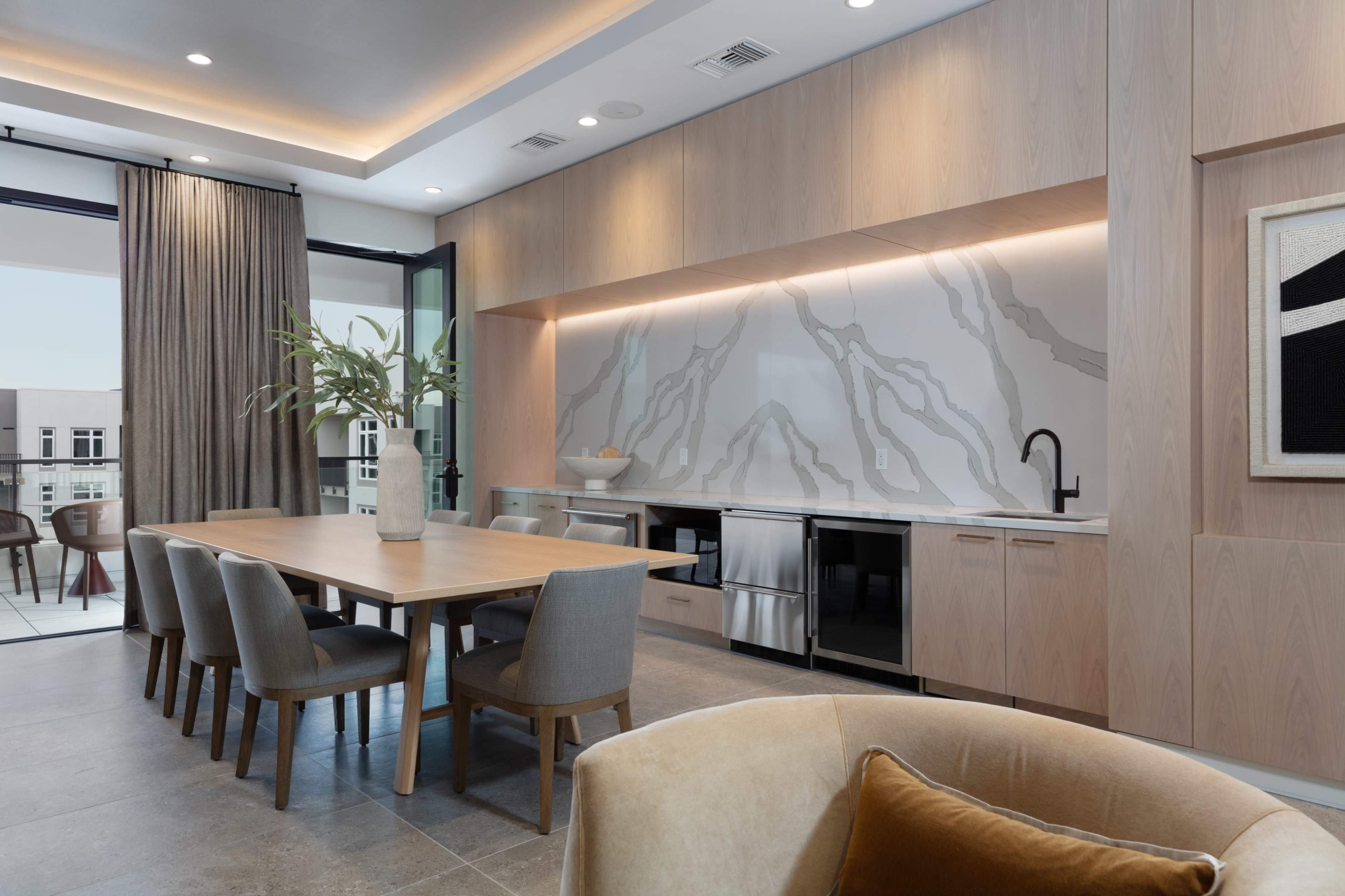 The image depicts a modern dining area with a wooden table surrounded by upholstered chairs, an elegant countertop with a marble backsplash, and large windows allowing natural light to enter.