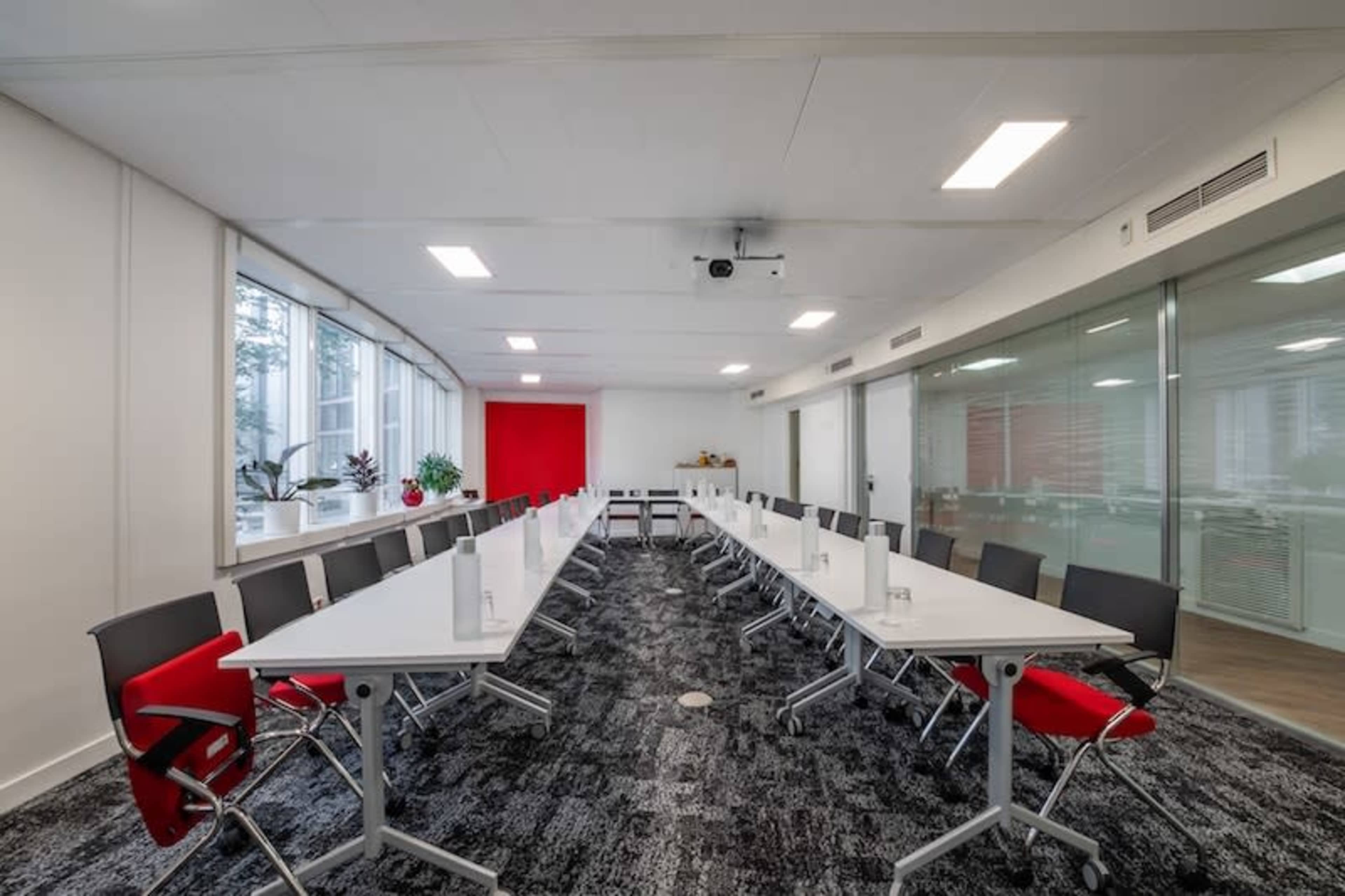 A spacious conference room features a long arrangement of tables and chairs, large windows, and a red accent wall.