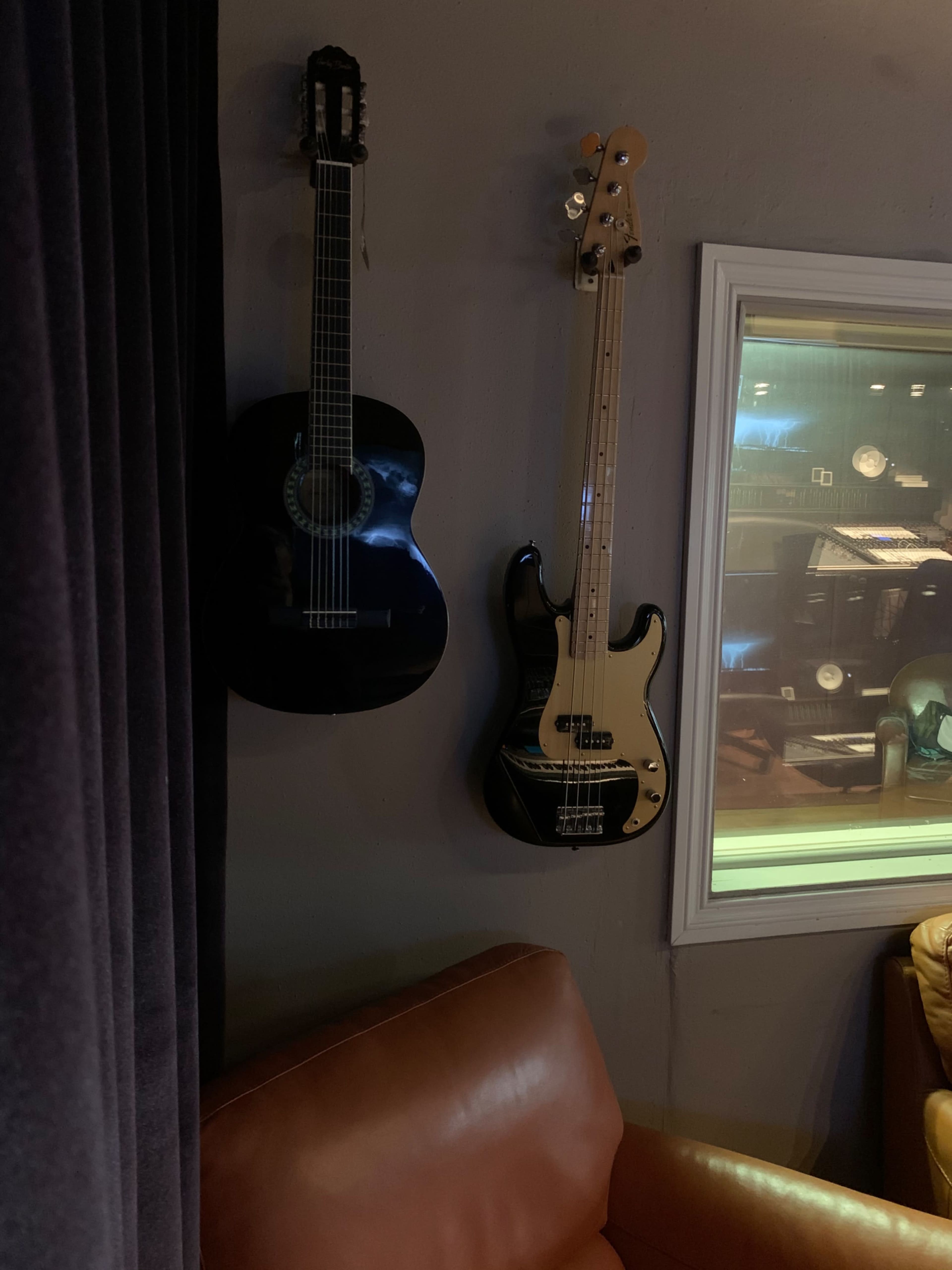 A black acoustic guitar and a black electric bass guitar hang on a gray wall beside a brown leather sofa.