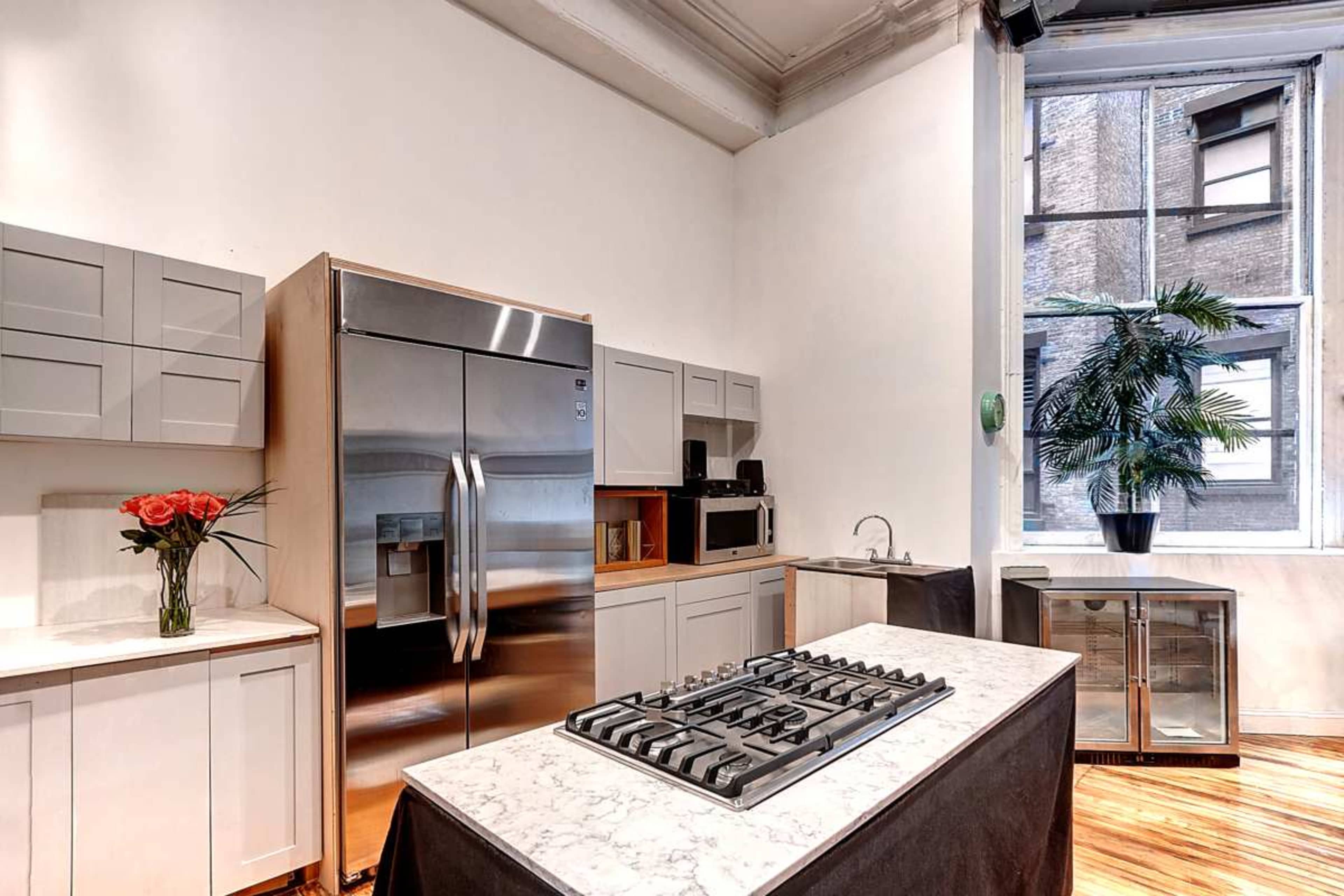 The image shows a modern kitchen featuring stainless steel appliances, light gray cabinetry, and a marble countertop with a gas stove in the center, along with a small dining area and a large window.