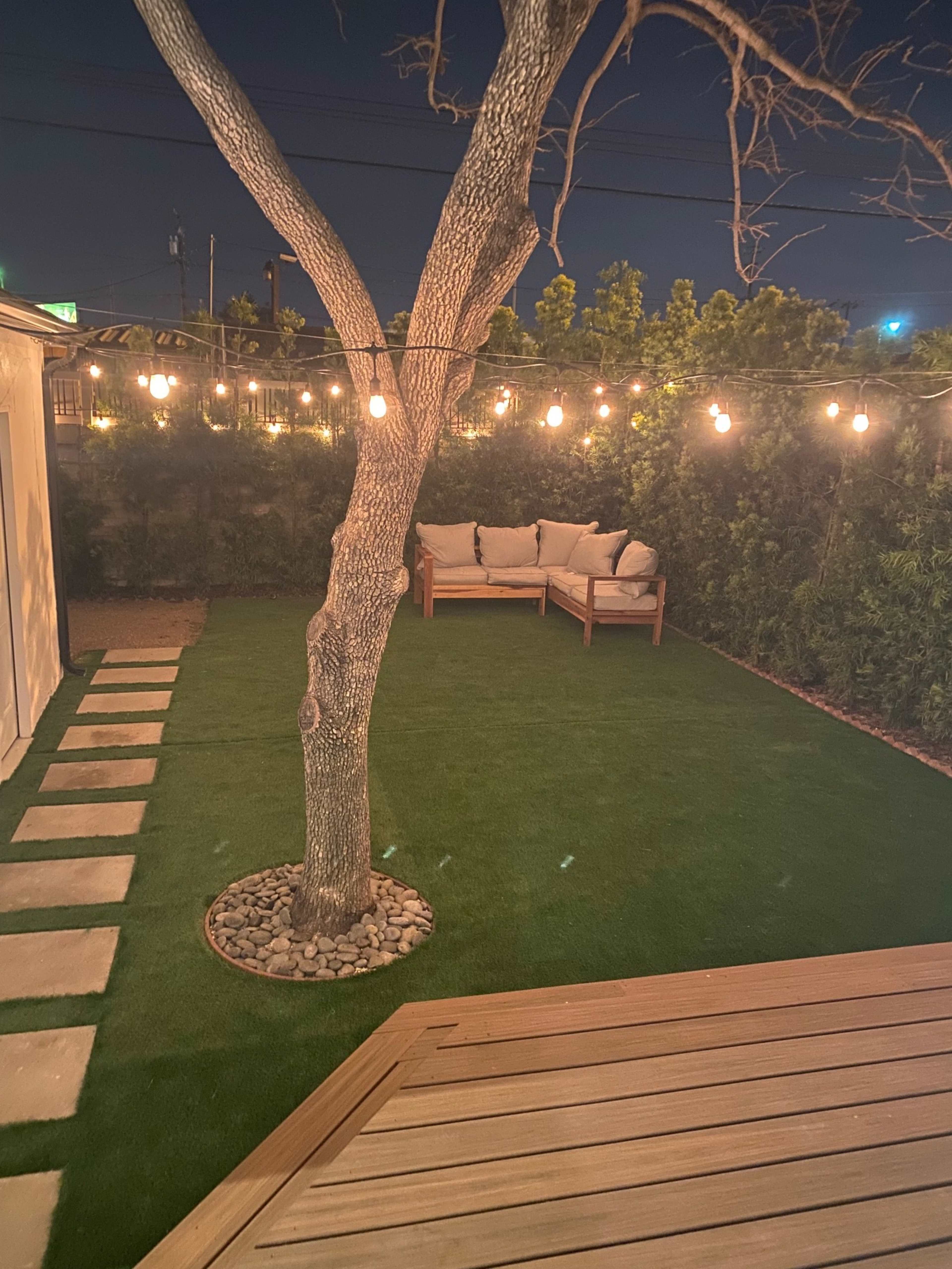 The image shows a backyard area at night, featuring a grassy section with a tree, a seating arrangement, stone pathways, and string lights overhead.