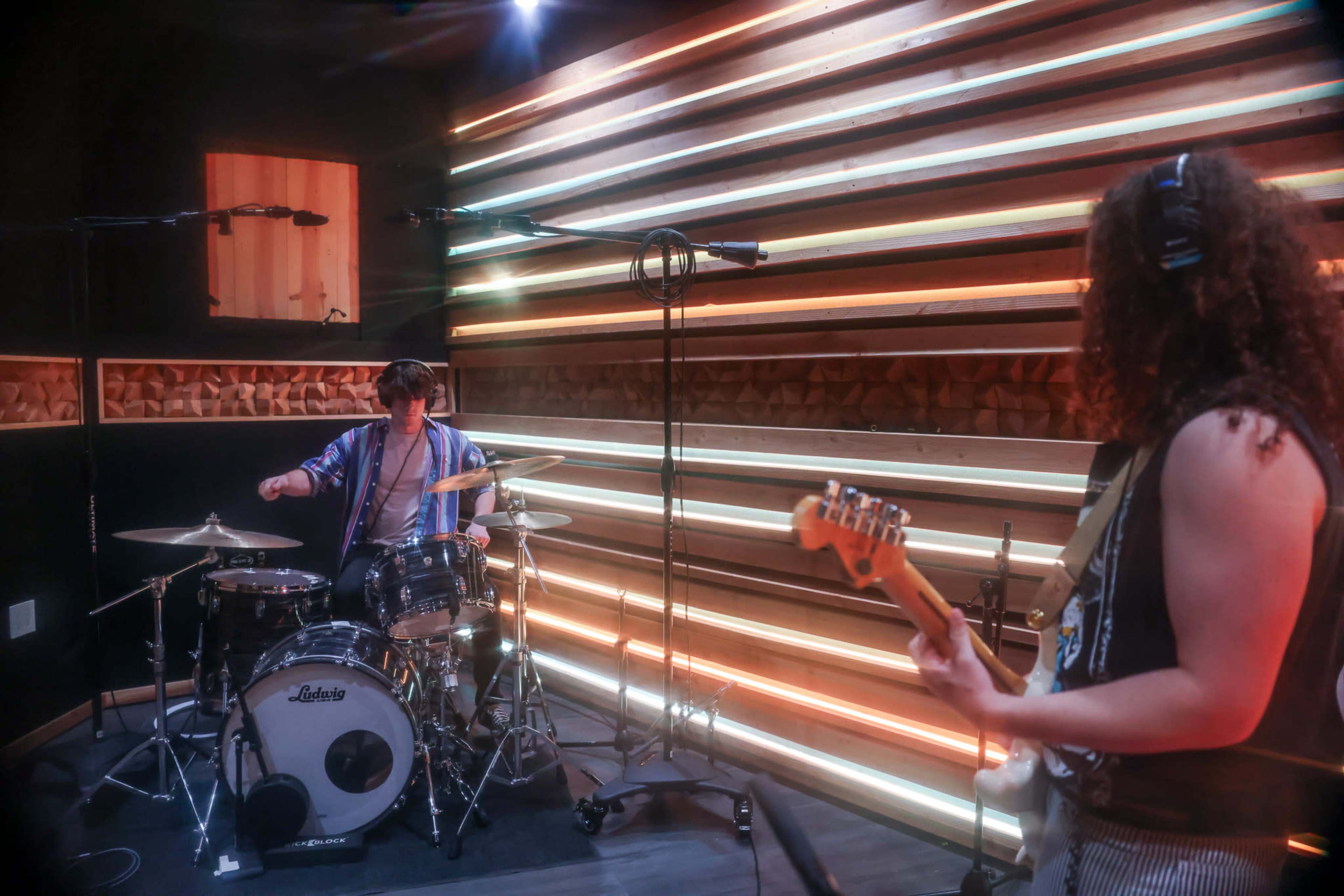 Two musicians are performing in a recording studio, with one playing drums and the other playing an electric bass guitar against a backdrop of illuminated wooden walls.