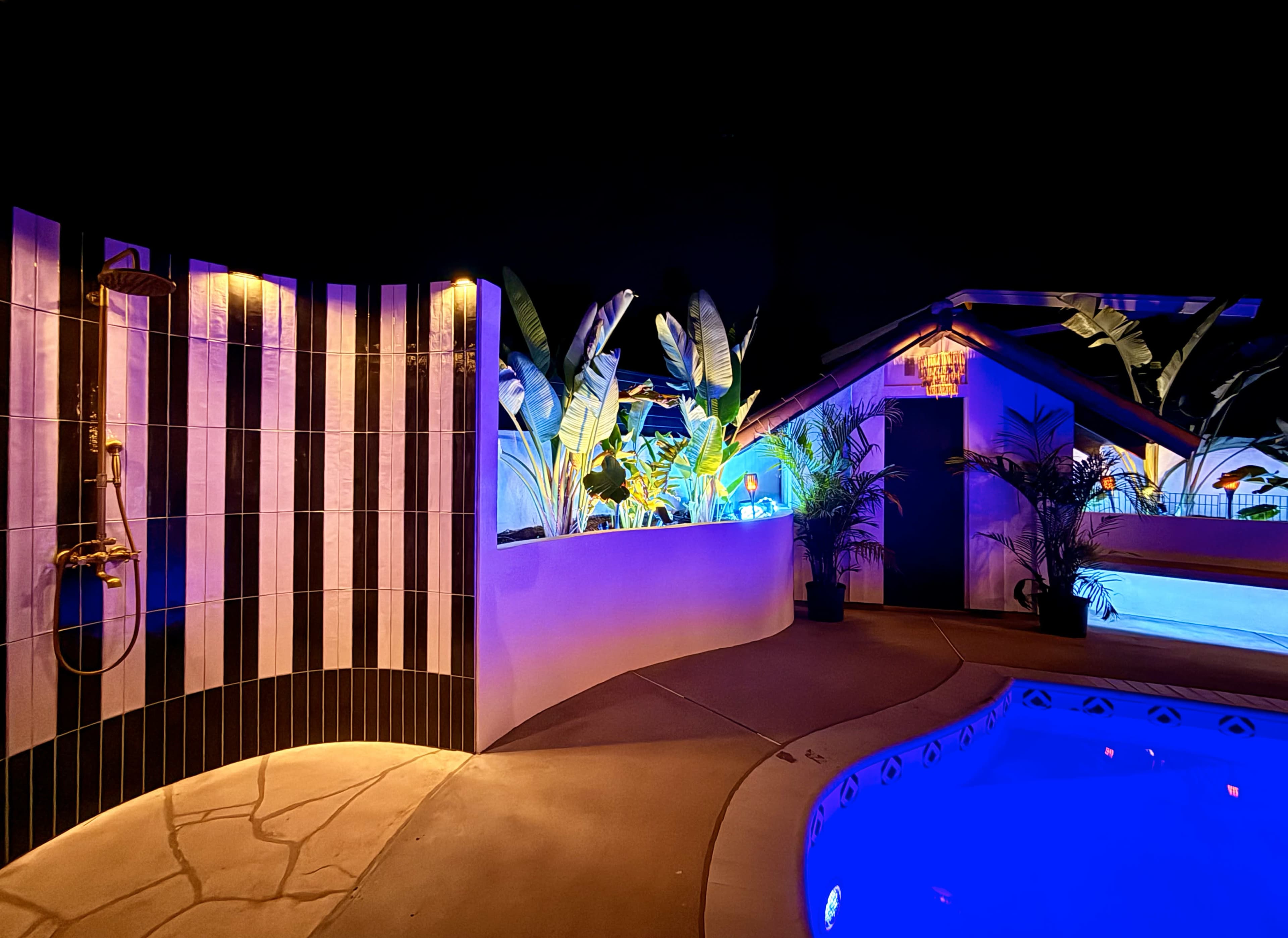 The image shows a well-lit outdoor area featuring a swimming pool, a shower with a striped wall, and tropical plants illuminated by colorful lights.