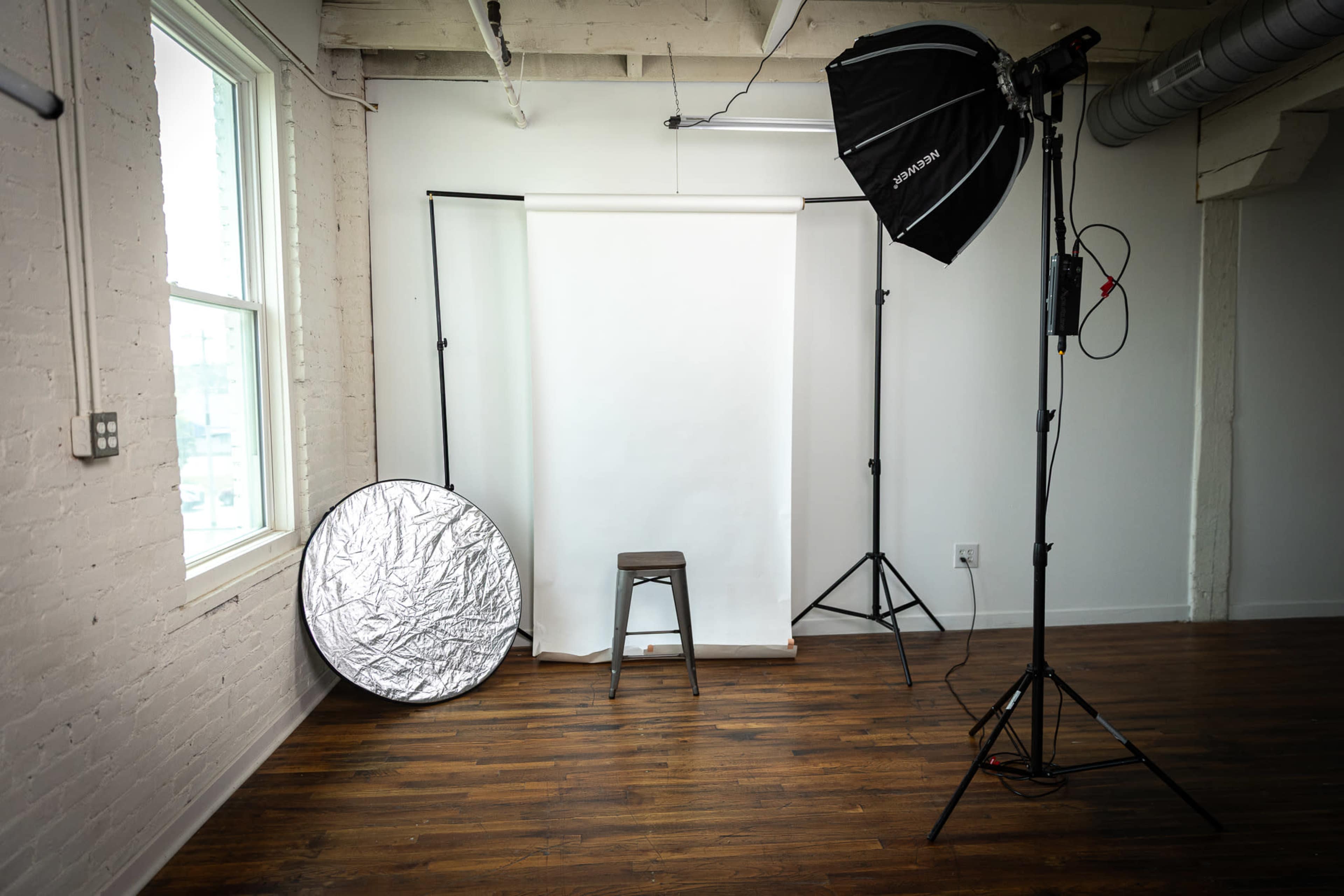 The image shows a photography studio setup with a white backdrop, a stool, a reflector, and studio lights against a wall with a window.