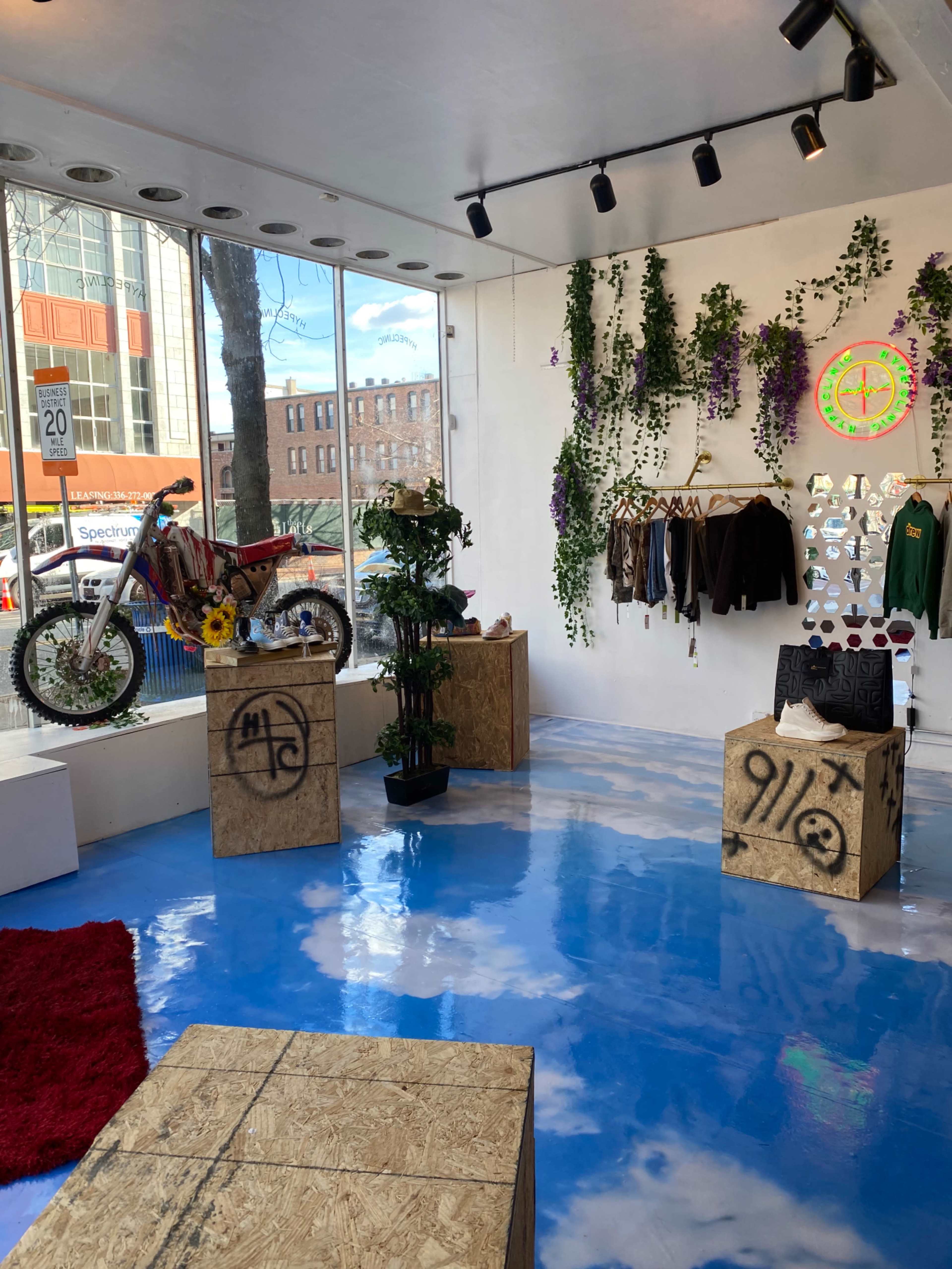 The interior of a contemporary space features a dirt bike on a wall, clothing displayed on hangers, greenery along the walls, and wooden boxes as tables on a blue floor that resembles a cloudy sky.